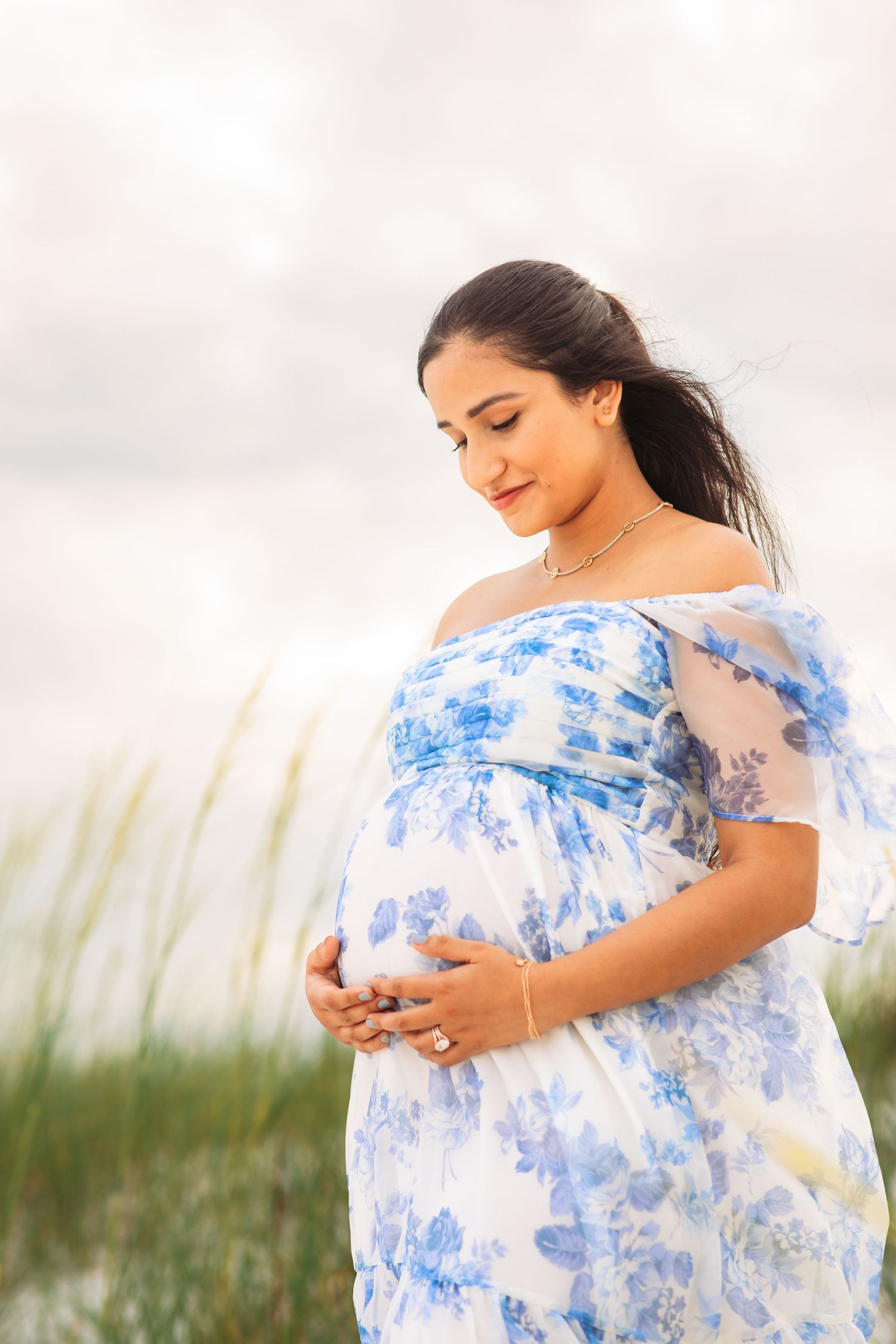 Expectant mother in blue floral dress poses peacefully near beach grass.