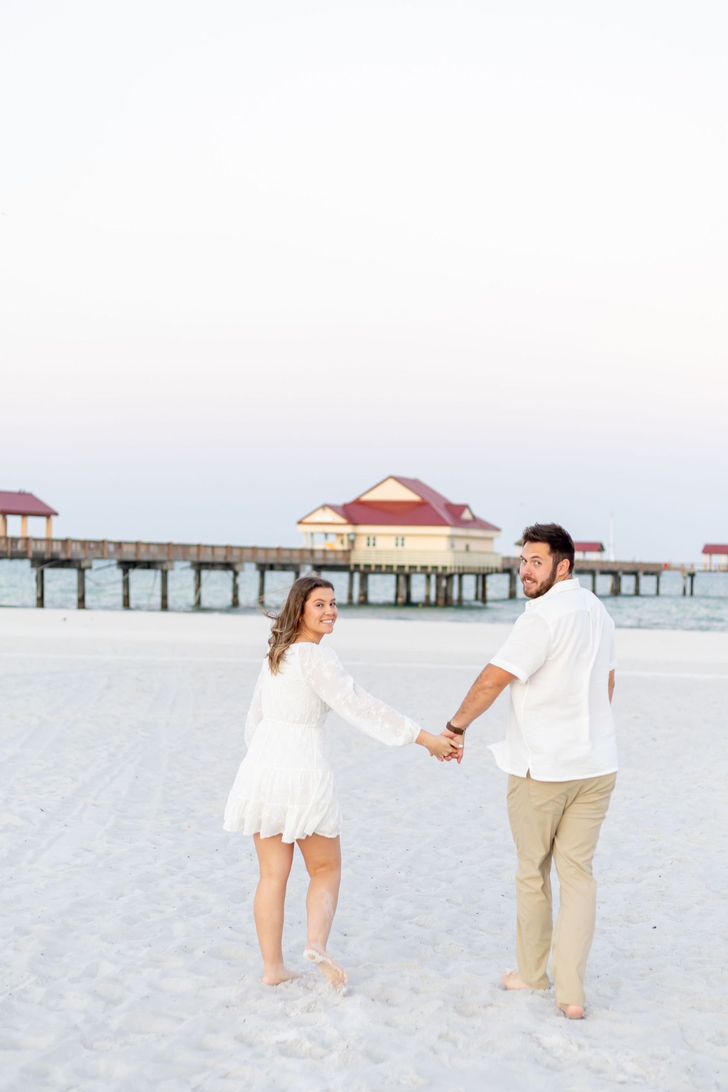 Couple holding hands on white sand beach near pier at sunset, woman in white dress, man in white shirt and khakis.
