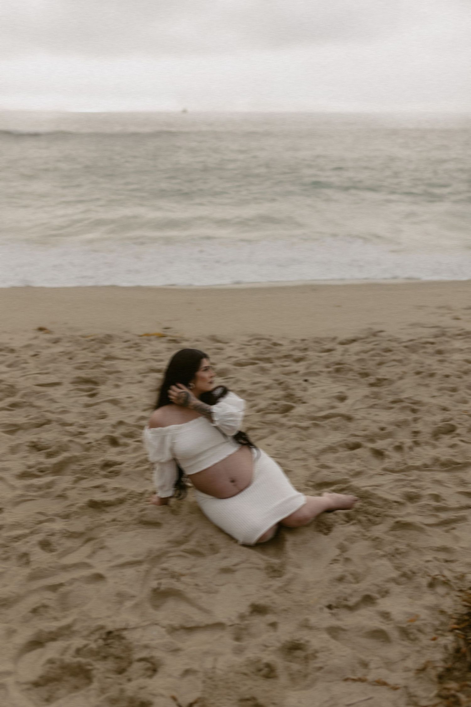 Expecting mother in white dress sits on cloudy beach during maternity photoshoot.