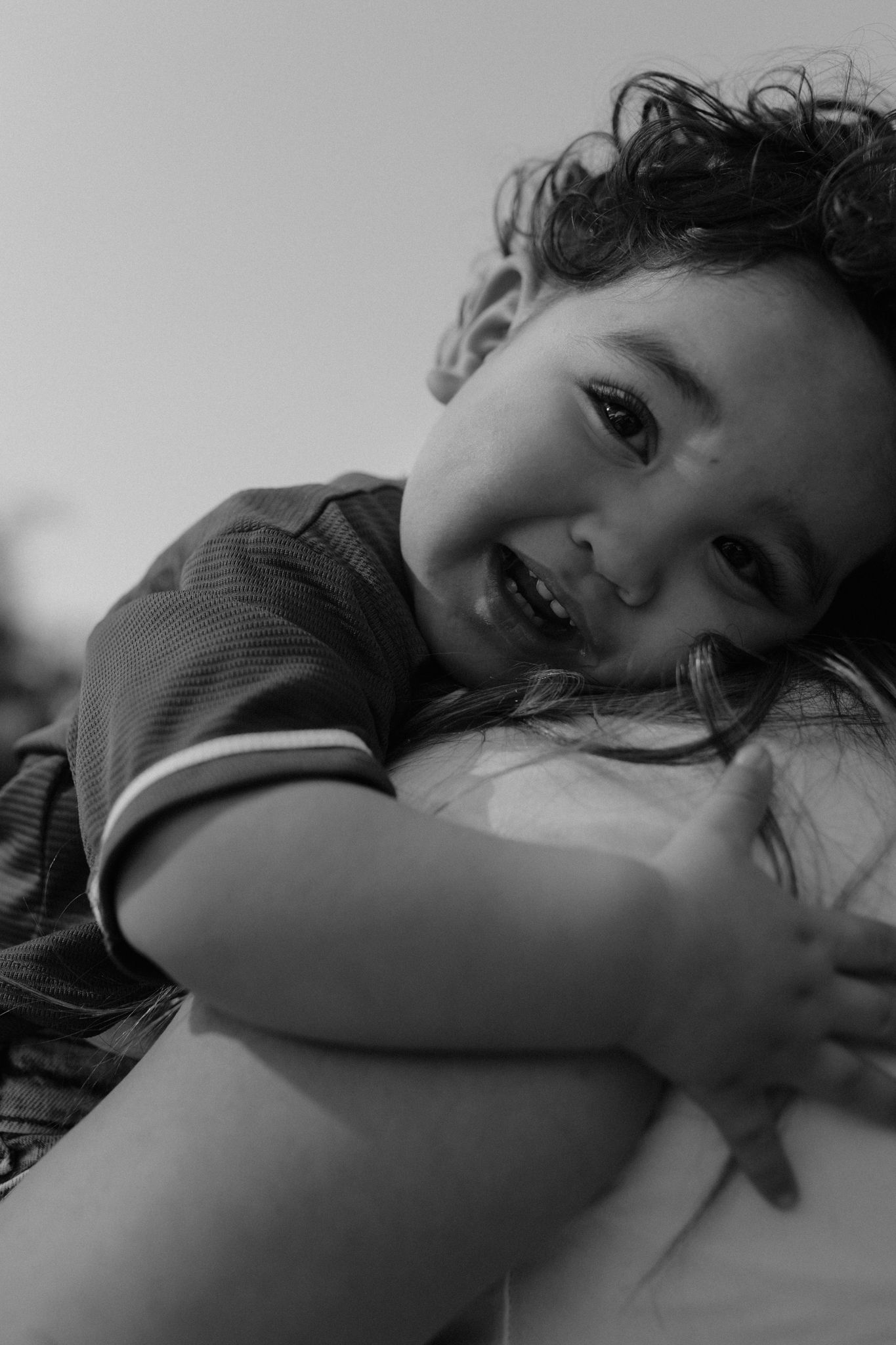 Black and white portrait of child with curly hair smiling while hugging someone in candid joyful moment.