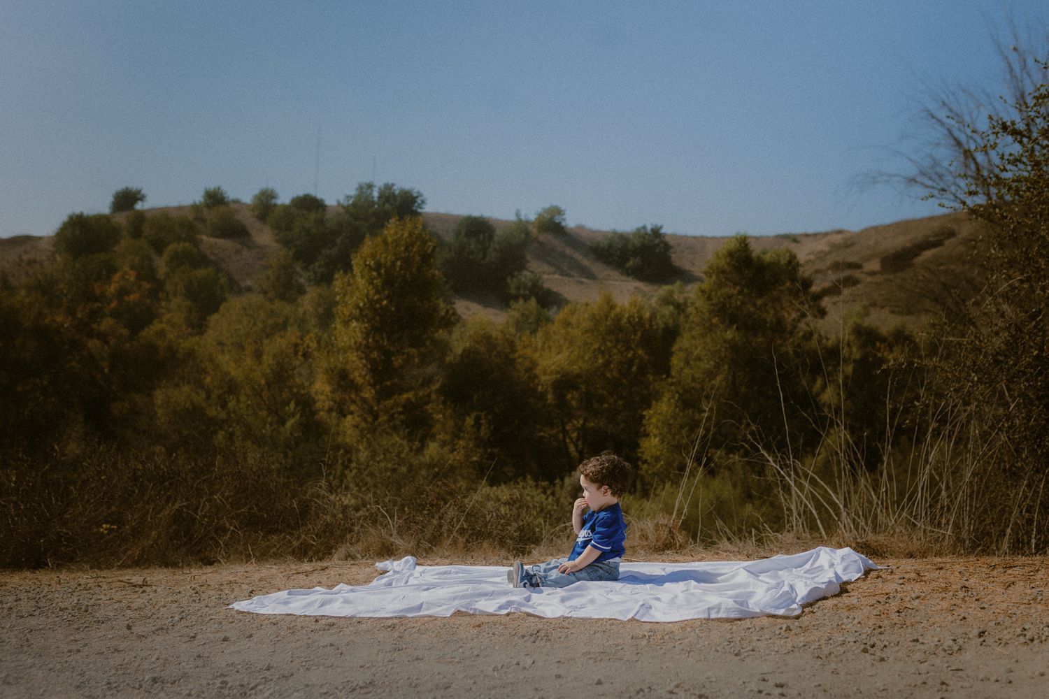 A small figure sits on a white blanket on sandy ground with autumn trees and rolling hills in the background.