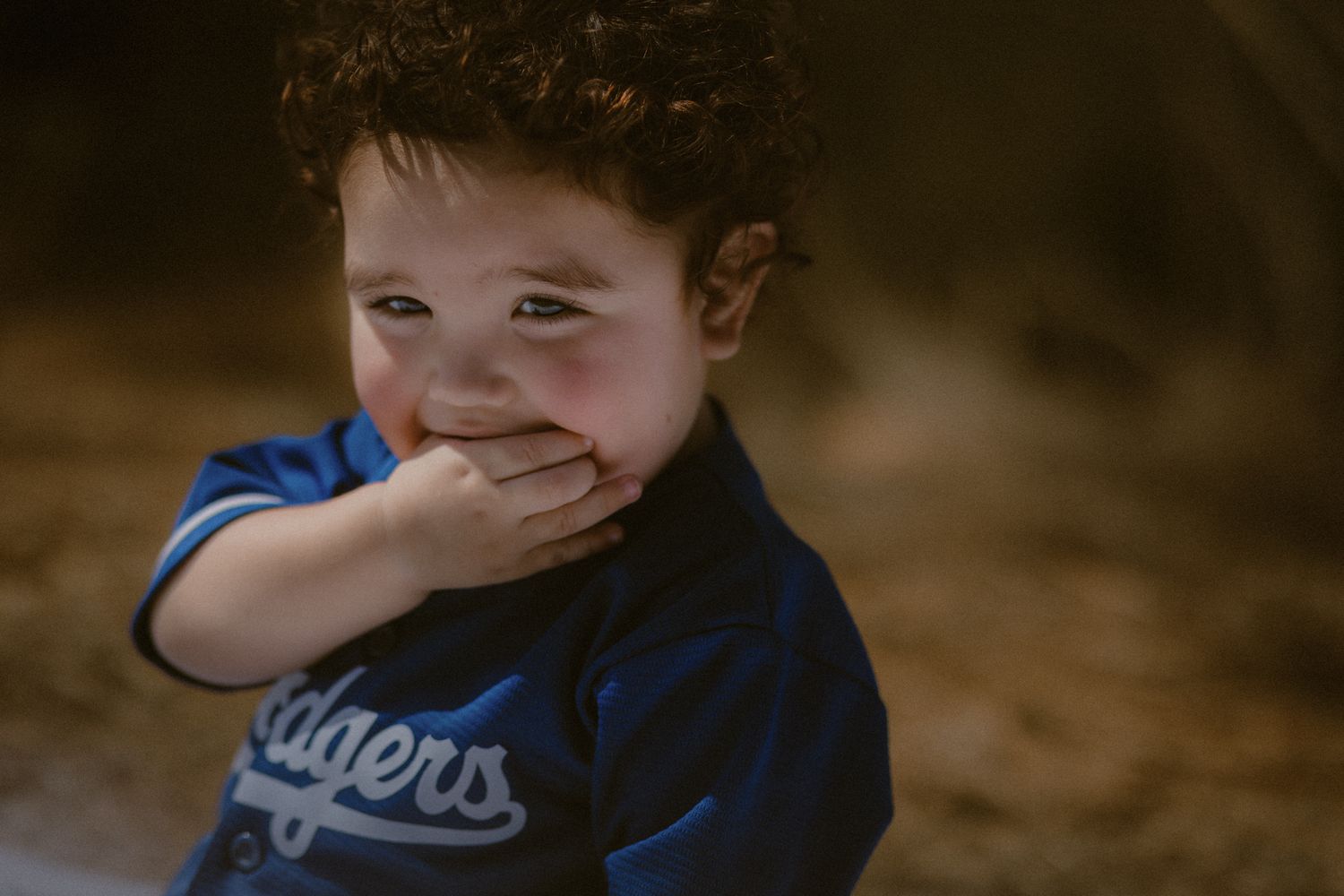 A child in a blue Dodgers shirt giggling with hand over their mouth against a blurred brown background.