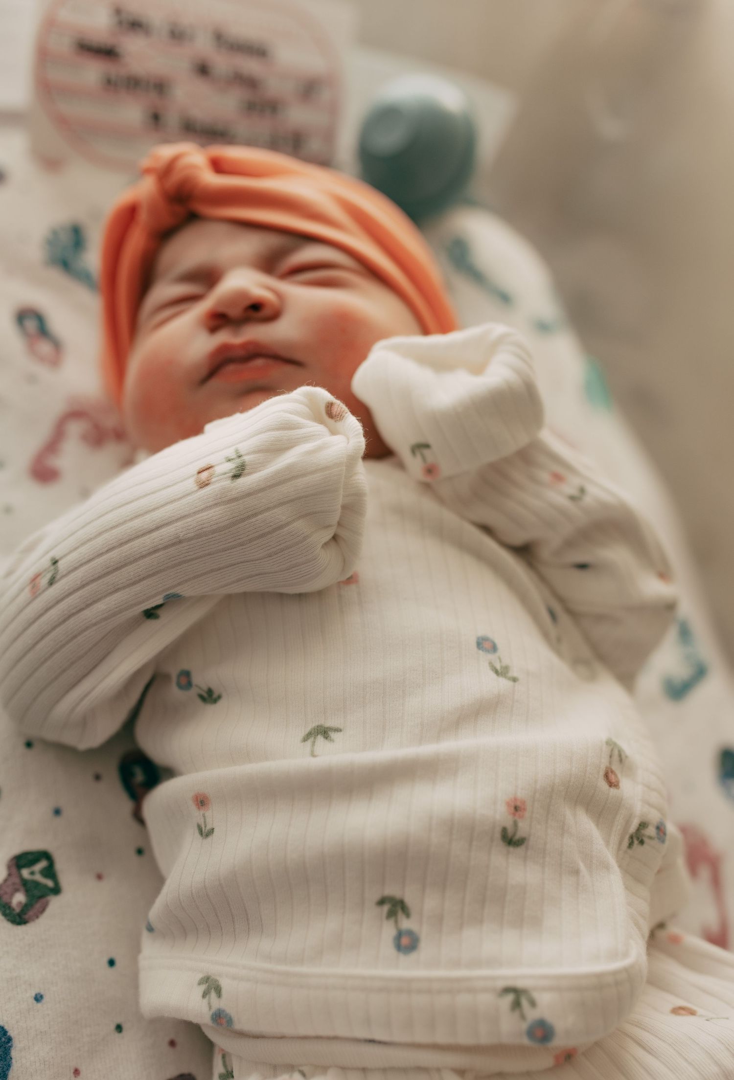 A sleeping newborn wearing a peach colored hat and white floral print onesie in the hospital.