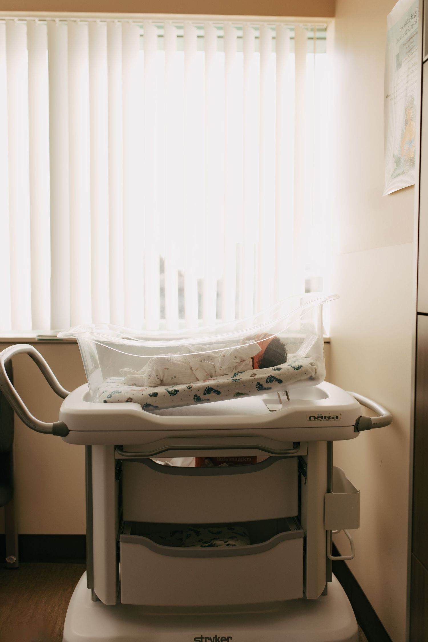 A medical examination bed with white linens sits next to vertical blinds in a clinical room setting.