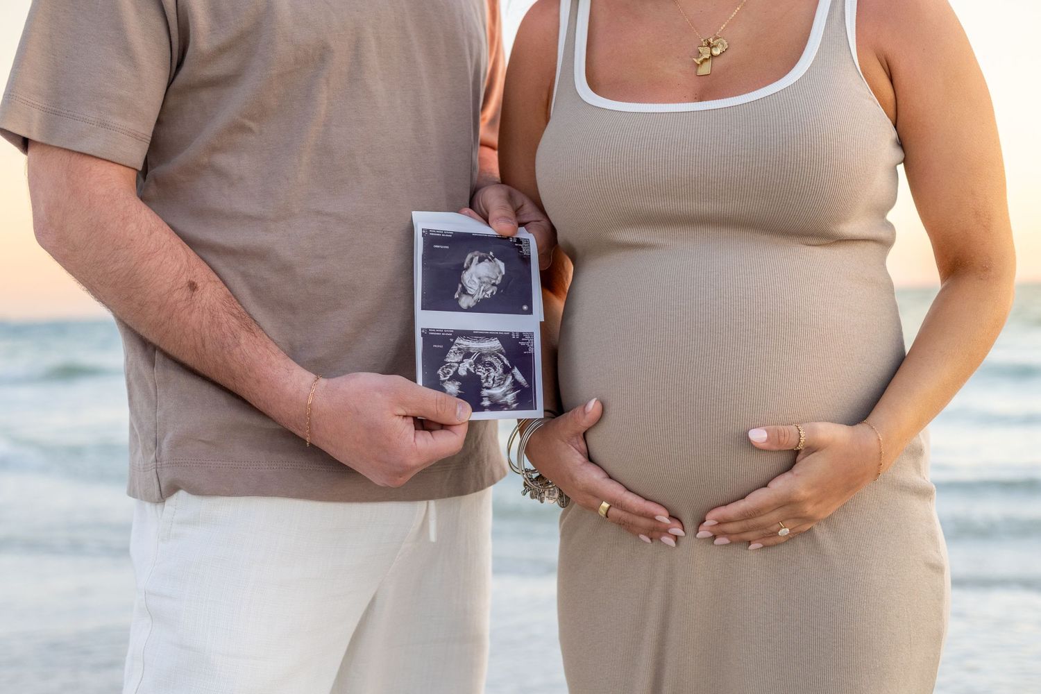 Expecting couple holding ultrasound photos in front of ocean during beach maternity photo session.
