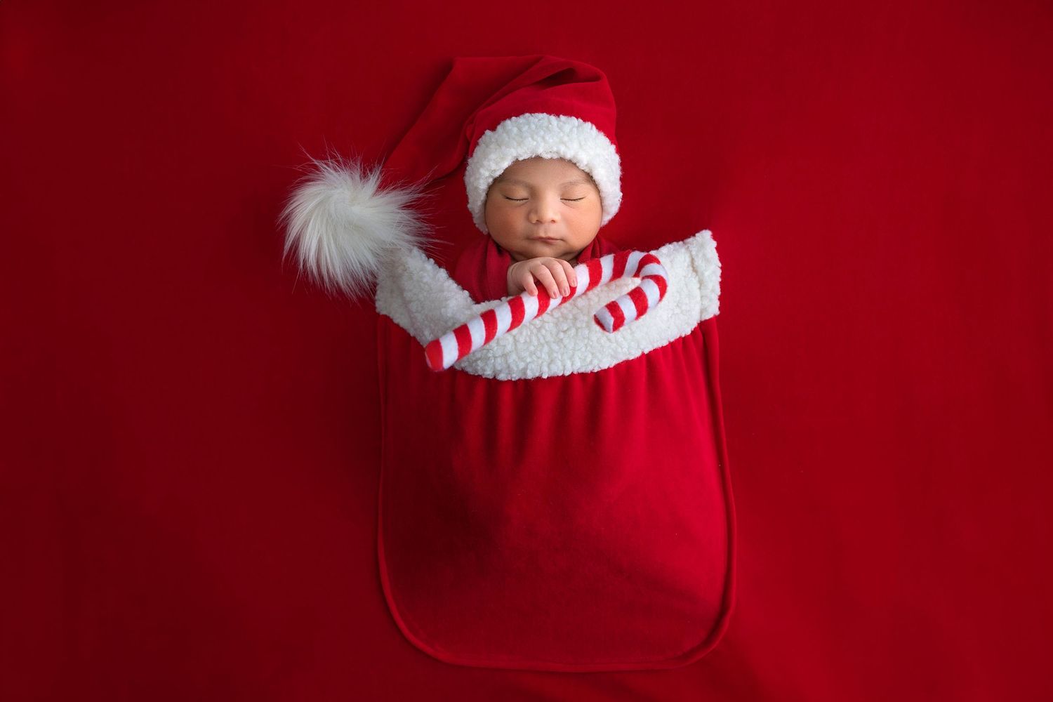 A newborn baby wearing a striped hat sleeps inside a red Christmas stocking against a red backdrop.