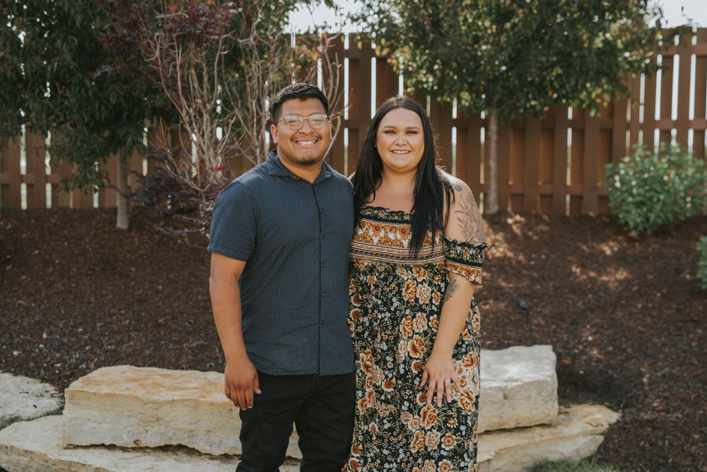Casual couple poses together in backyard setting with wooden fence.
