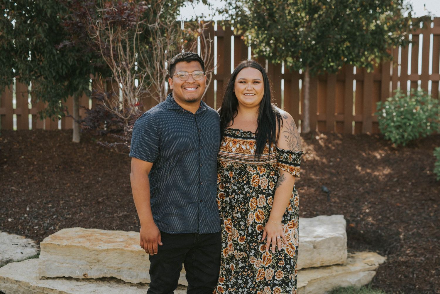 Casual couple poses together in backyard setting with wooden fence.
