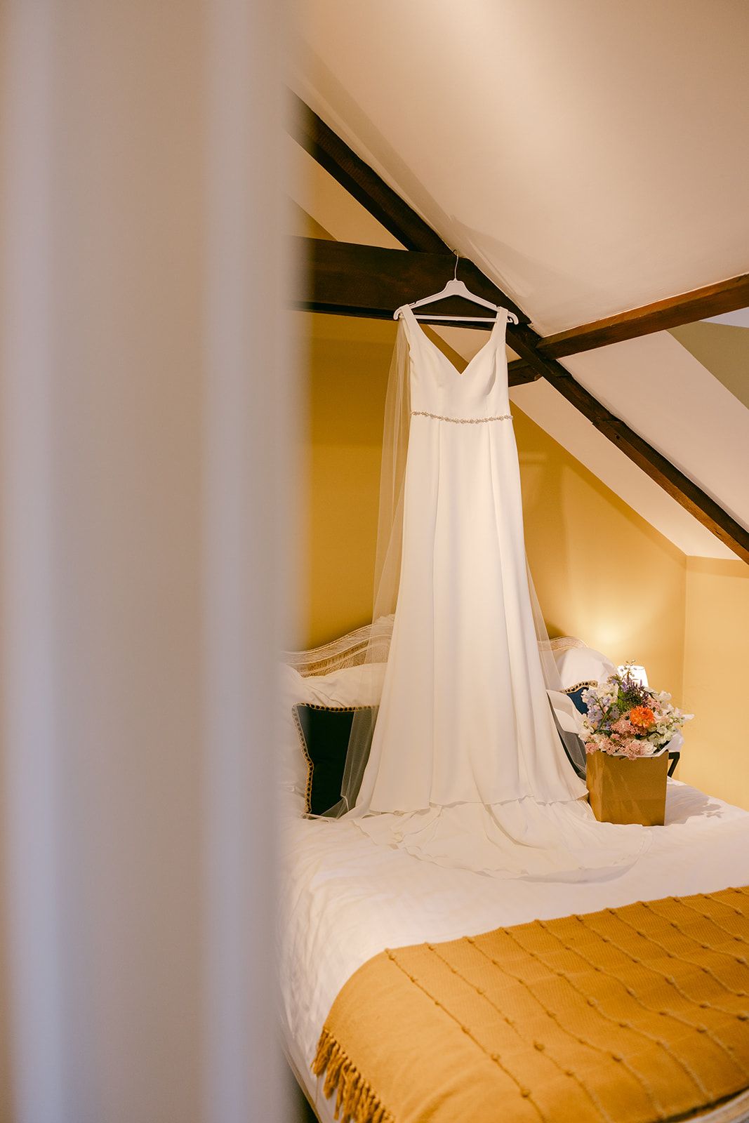 Elegant white wedding dress hanging from exposed wooden beam next to bridal bouquet in bridal cottage of Shortflatt Tower