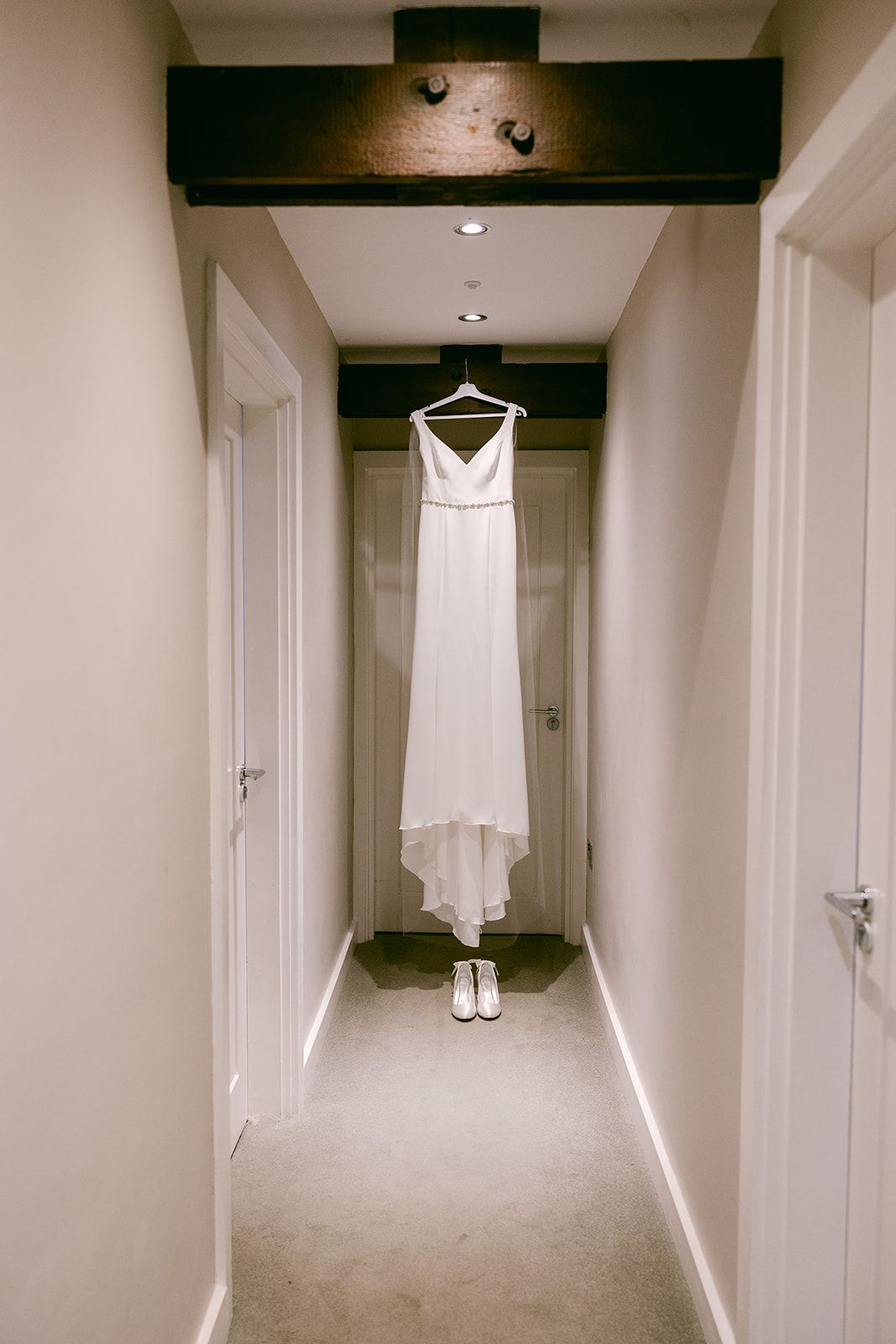 A white wedding dress and shoes hang elegantly in a hallway with wooden beams and white walls of bridal cottage at Shortflatt Tower