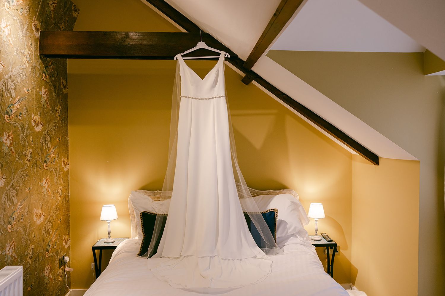 A simple white wedding dress hangs on display above a bed between two lit wall sconces in a golden-toned bedroom of Shortflatt Tower
