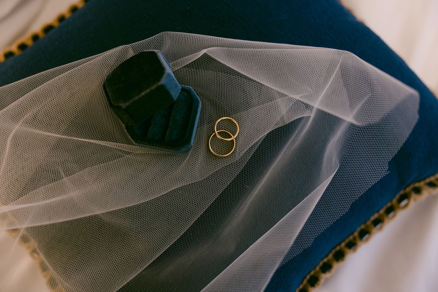 Two gold wedding rings rest on delicate white tulle veil fabric against a dark blue background.