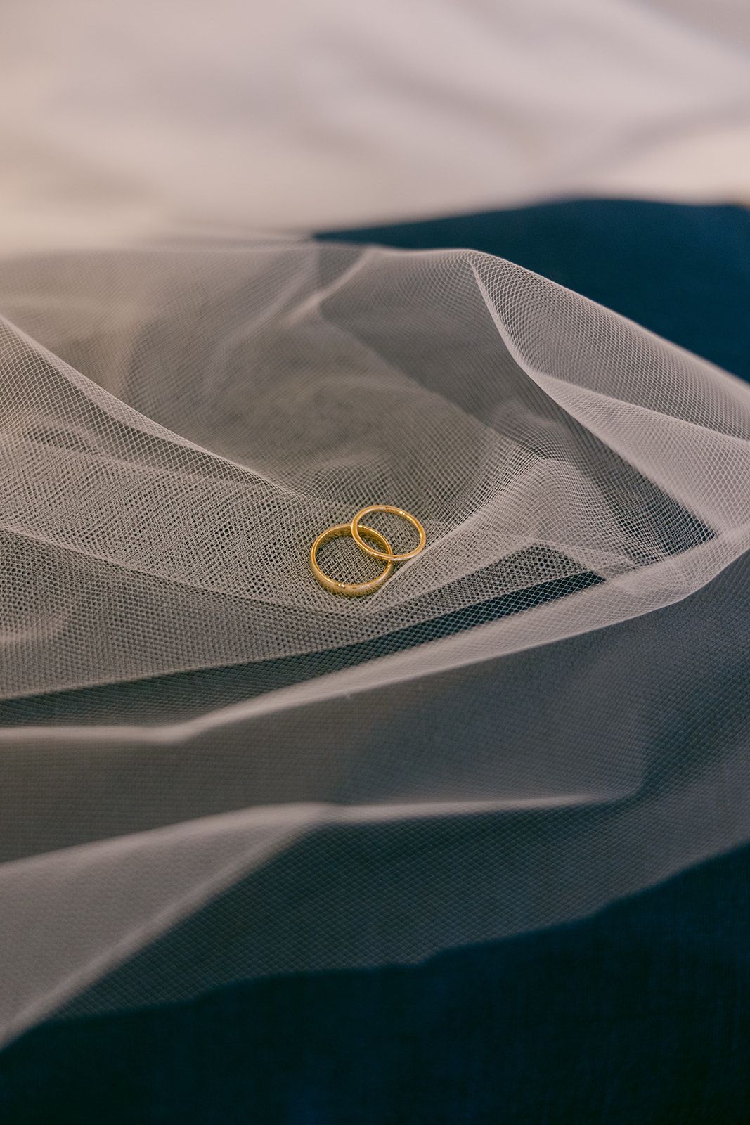 Two gold wedding rings rest delicately on flowing white tulle fabric with a dark background creating an elegant bridal detail.