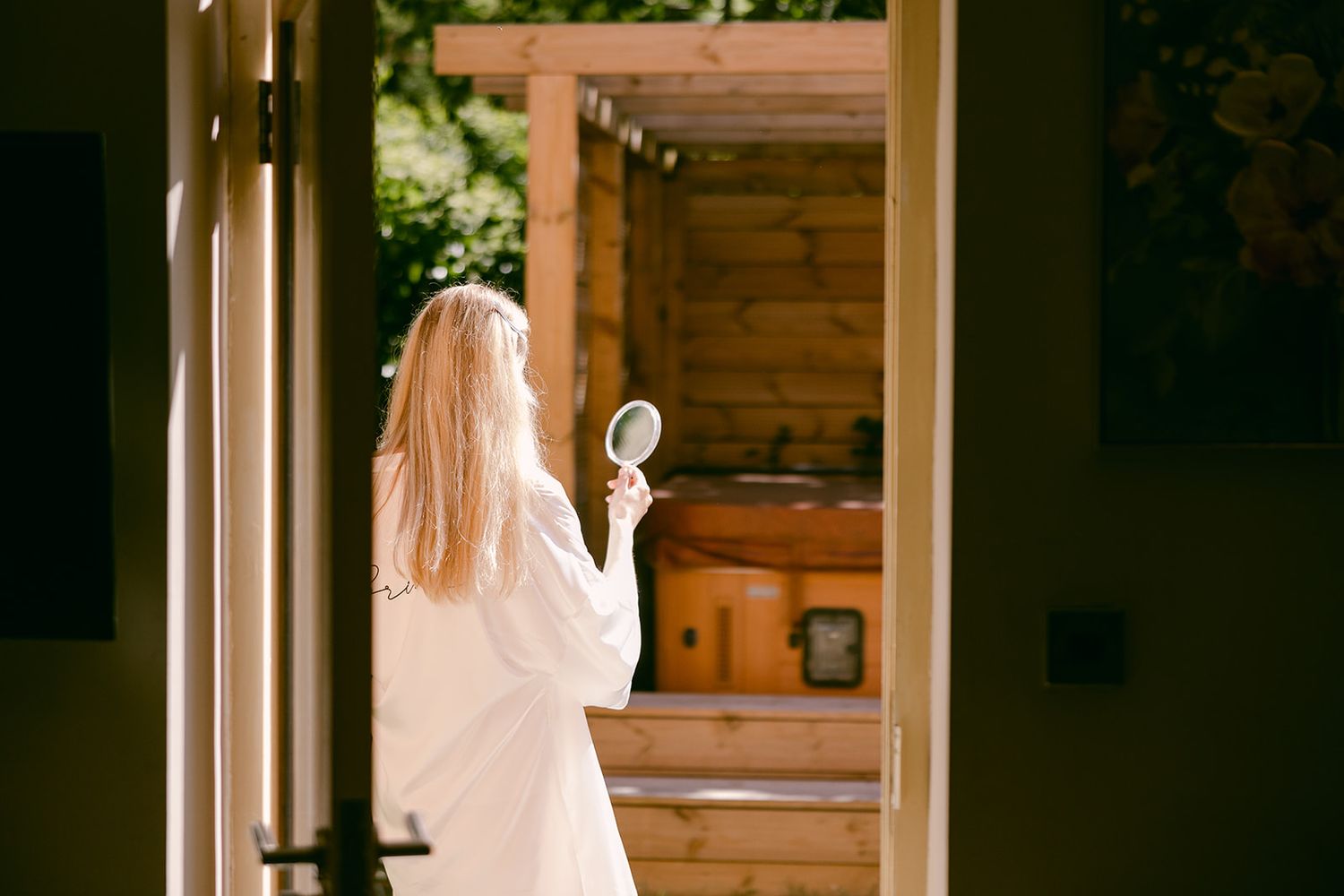 Bride with long blonde hair stands in doorway holding a white handheld mirror outside cottage at Shortflatt Tower