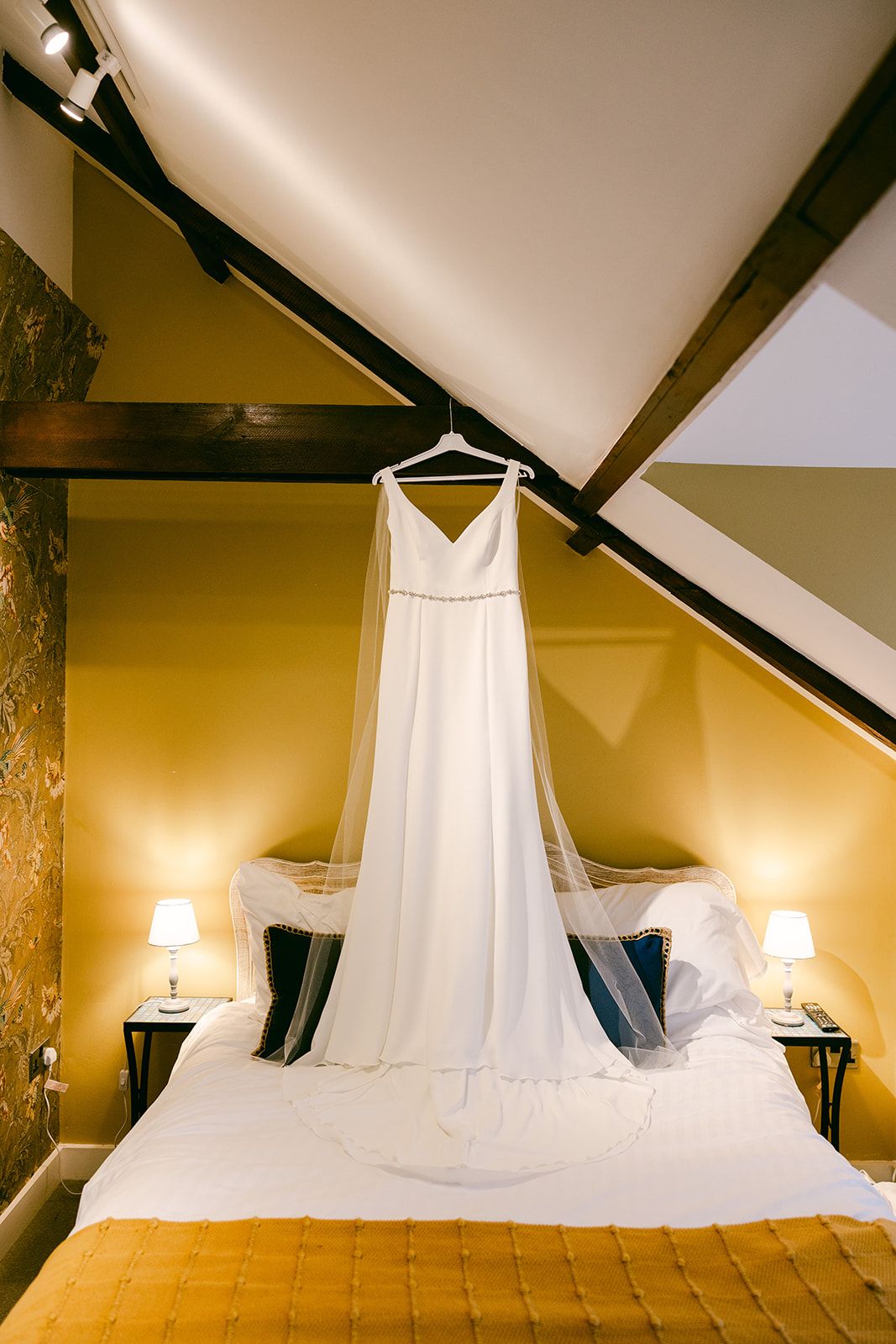 White wedding dress hangs elegantly over a golden bed in a cozy attic bedroom with warm lighting and exposed beams at Shortflatt Tower