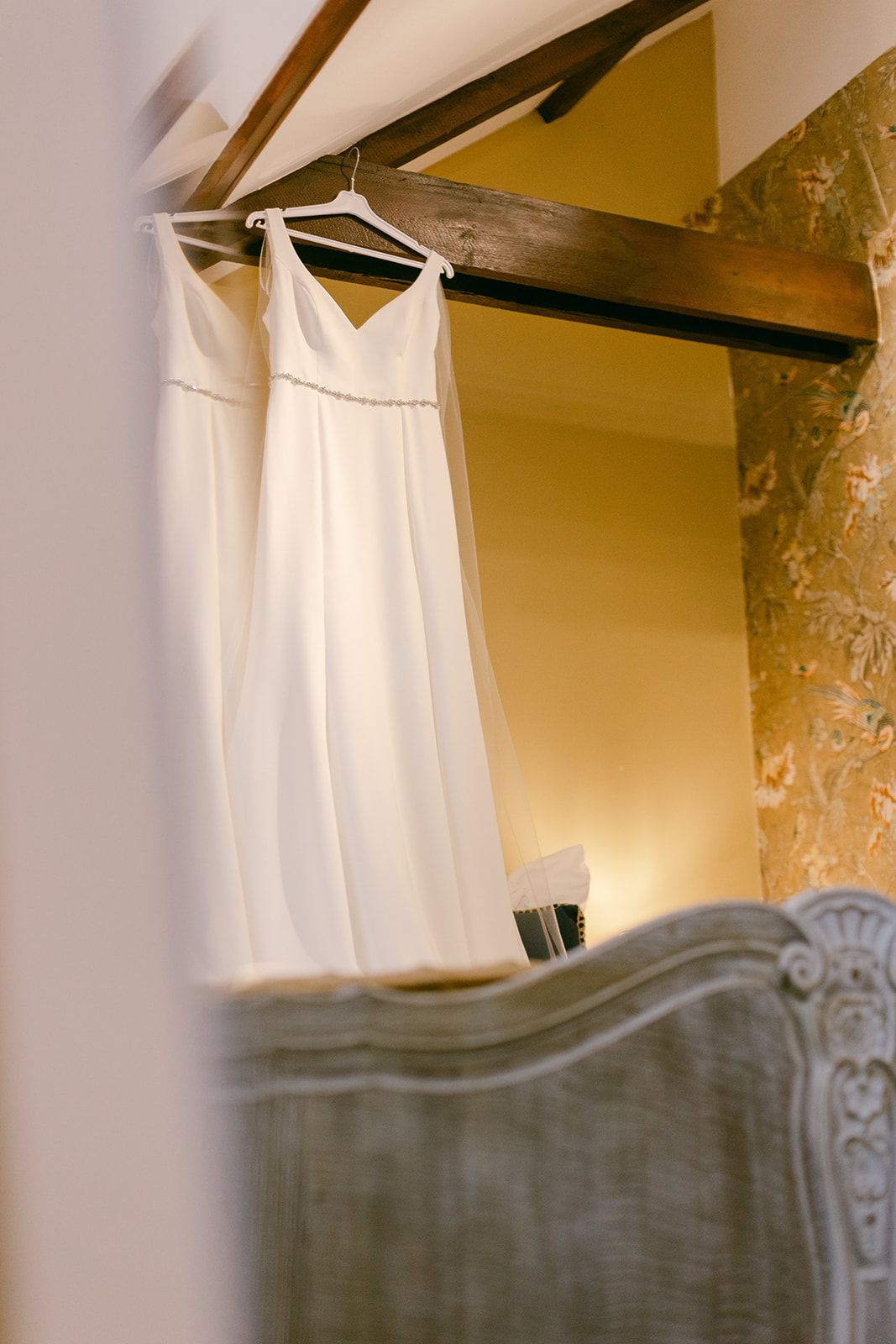 A simple white wedding dress hangs on a hanger against a yellow wall in bridal cottage of Shortflatt Tower Northumberland