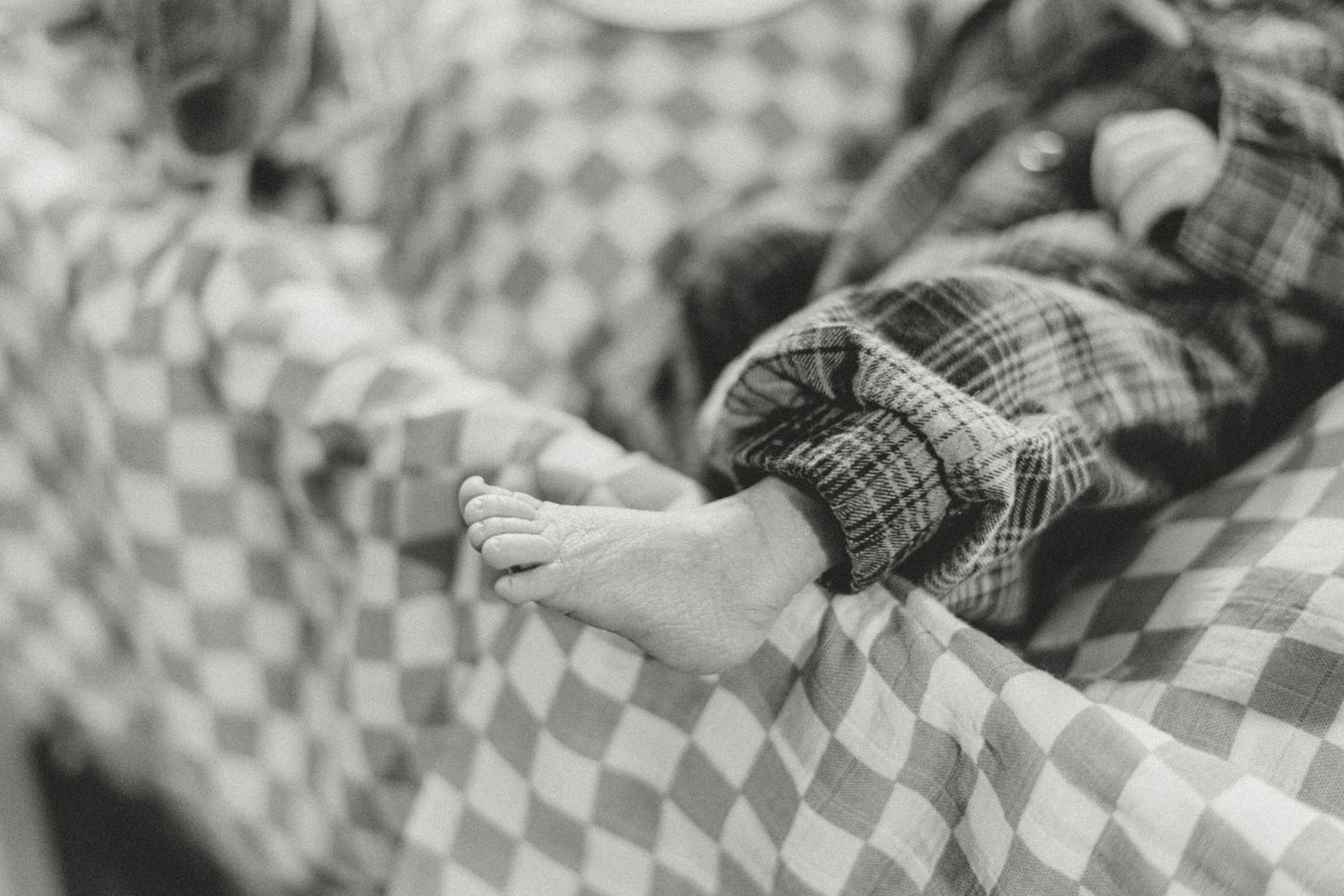 Close-up black and white photo of a baby's tiny foot resting on a checkered blanket pattern.