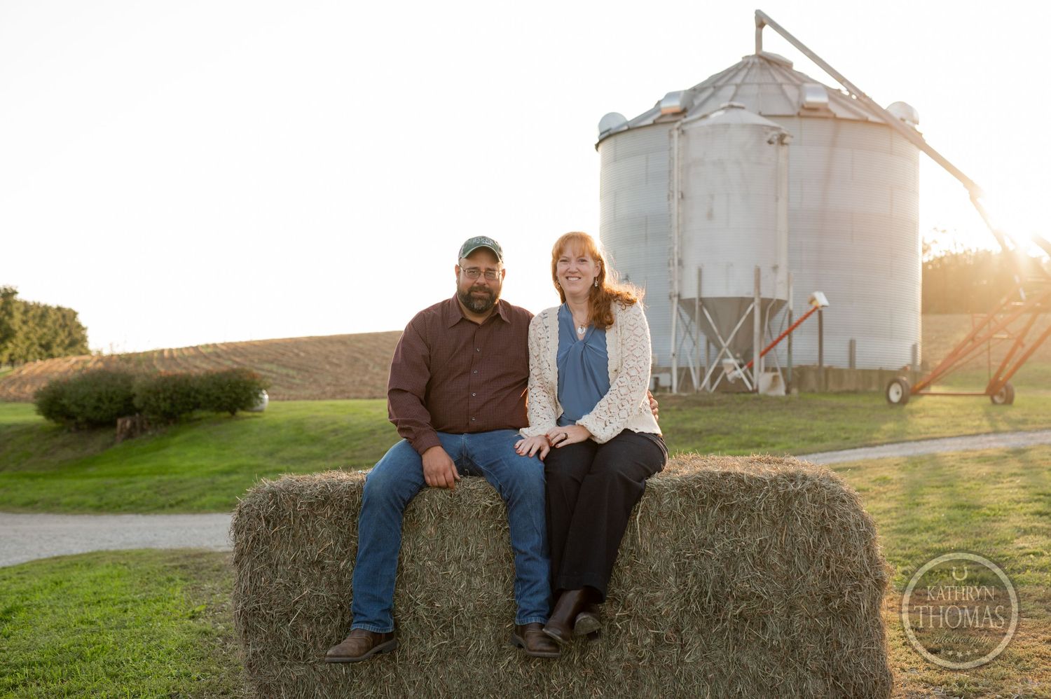 Couple sits on hay bale by grain silo during golden hour on farm.