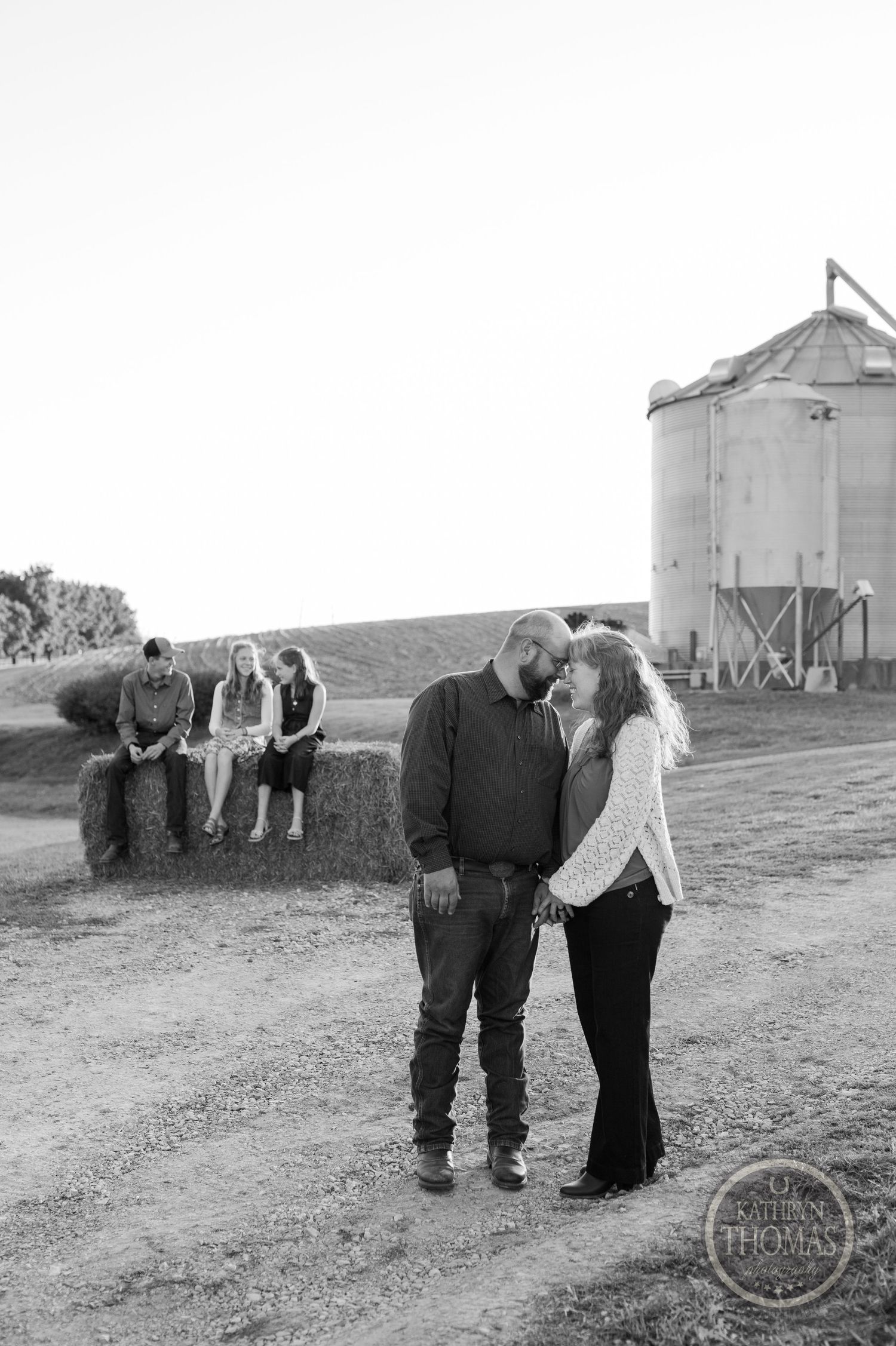 Black and white photo of couple in farm setting with grain silo and others in background.