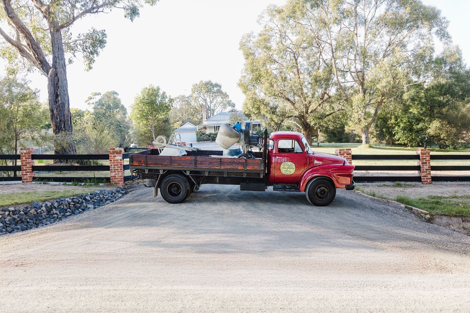 A vintage red flatbed truck parked on a gravel driveway with wooden fences and trees in the background.