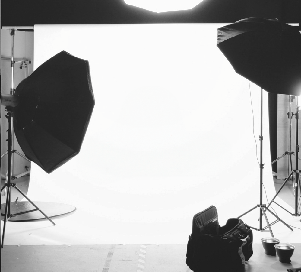 Professional photography studio setup with white backdrop, lighting umbrellas and equipment silhouetted against bright lights.
