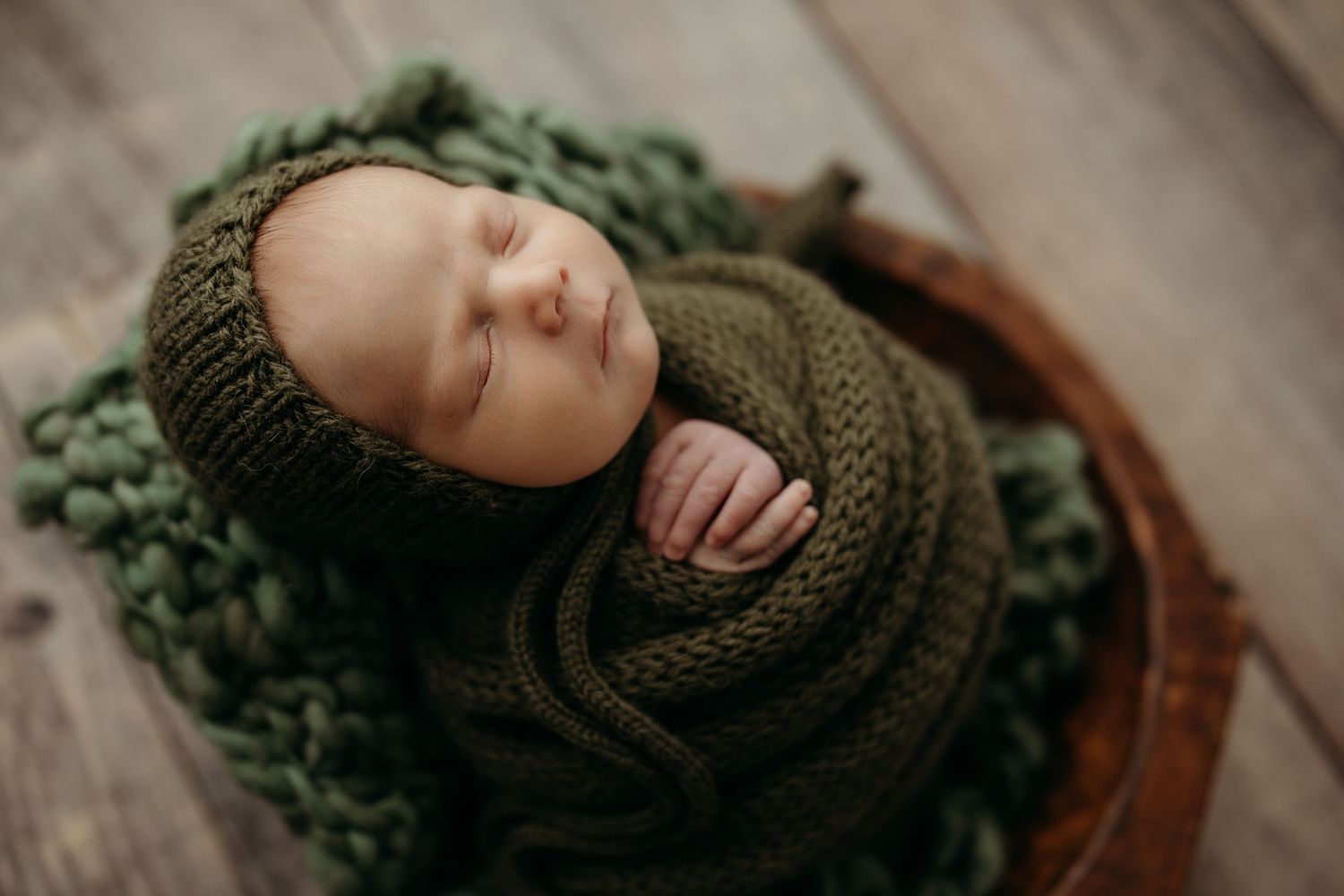 Close-up of sleeping baby wrapped in olive green knit wrap on wood background.