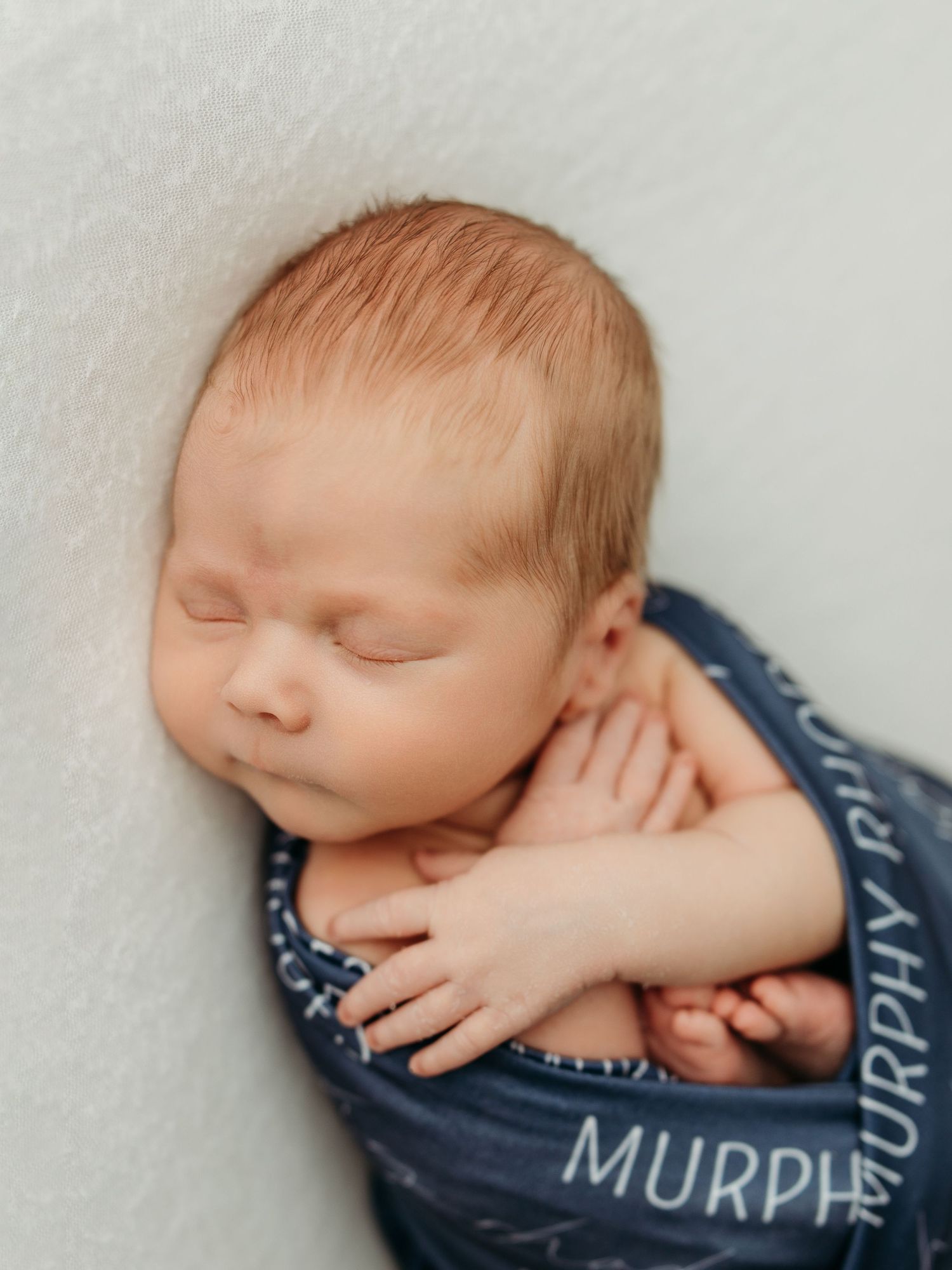 Peaceful newborn baby sleeping on a white blanket wearing a personalized navy blue wrap.