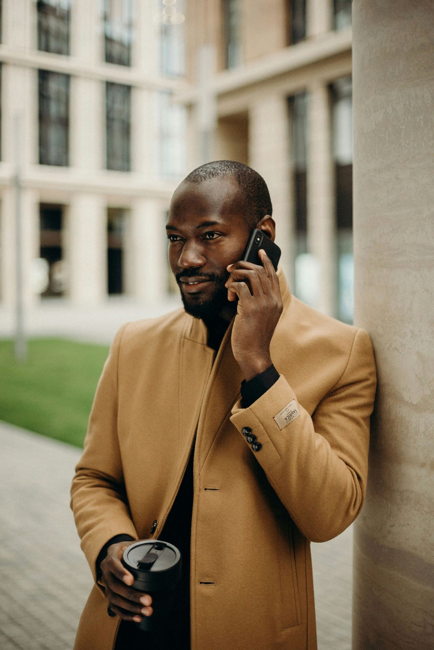Professional in camel coat talking on phone while holding coffee outside modern office building.