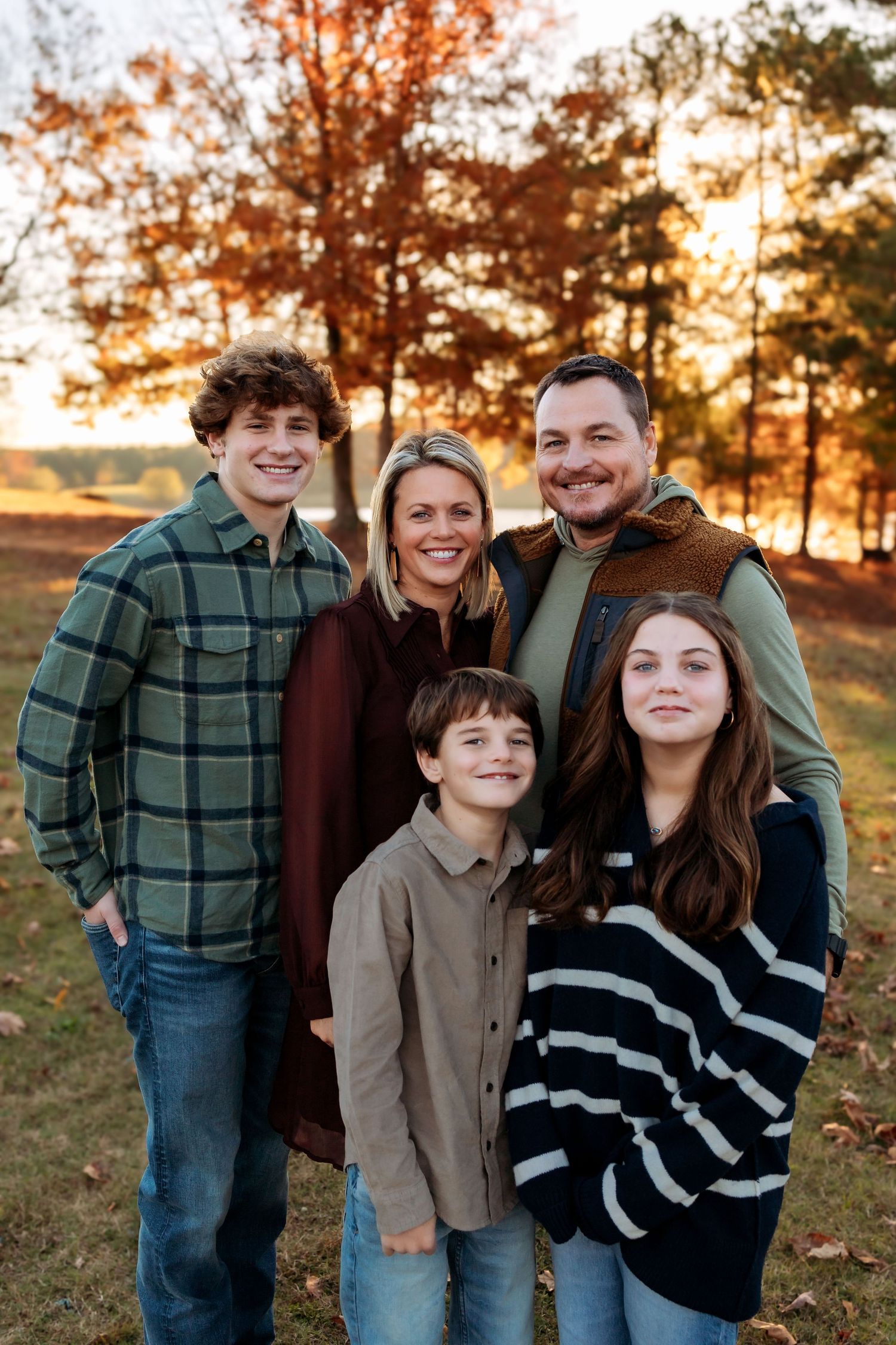 A family poses together on grass by a lake during golden hour sunset with trees in the background.