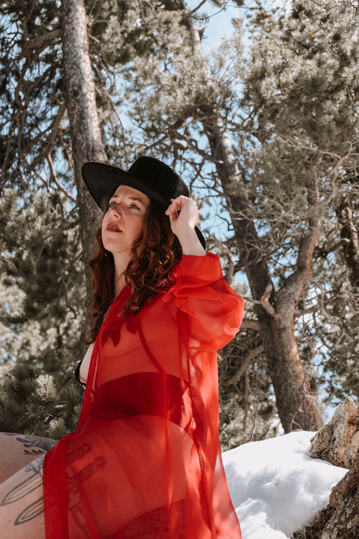 A woman in a bright red dress and black hat poses against a snowy winter landscape with pine trees in the background.