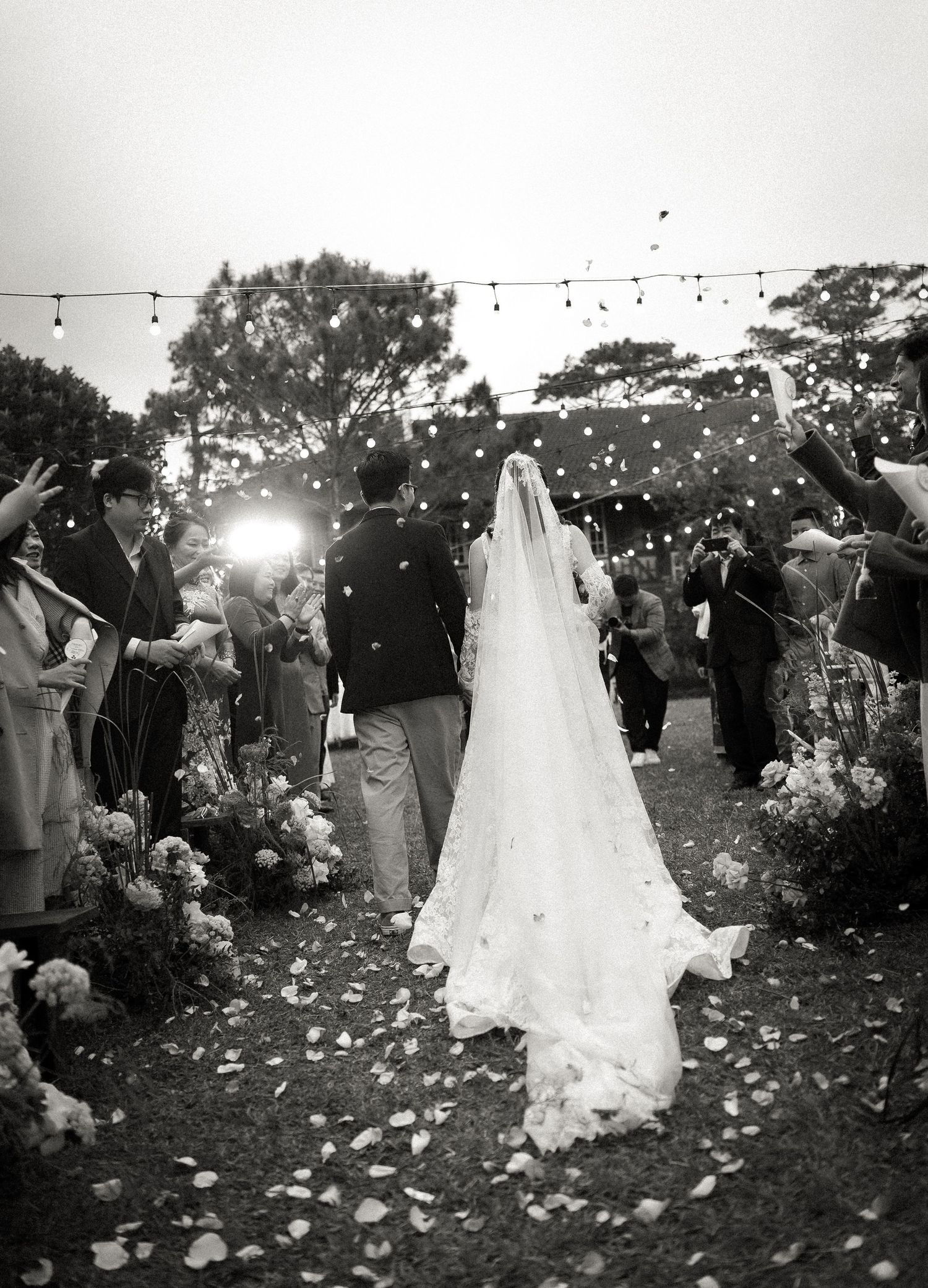 Black and white photo of an outdoor wedding ceremony with string lights overhead and flower petals scattered on the ground.