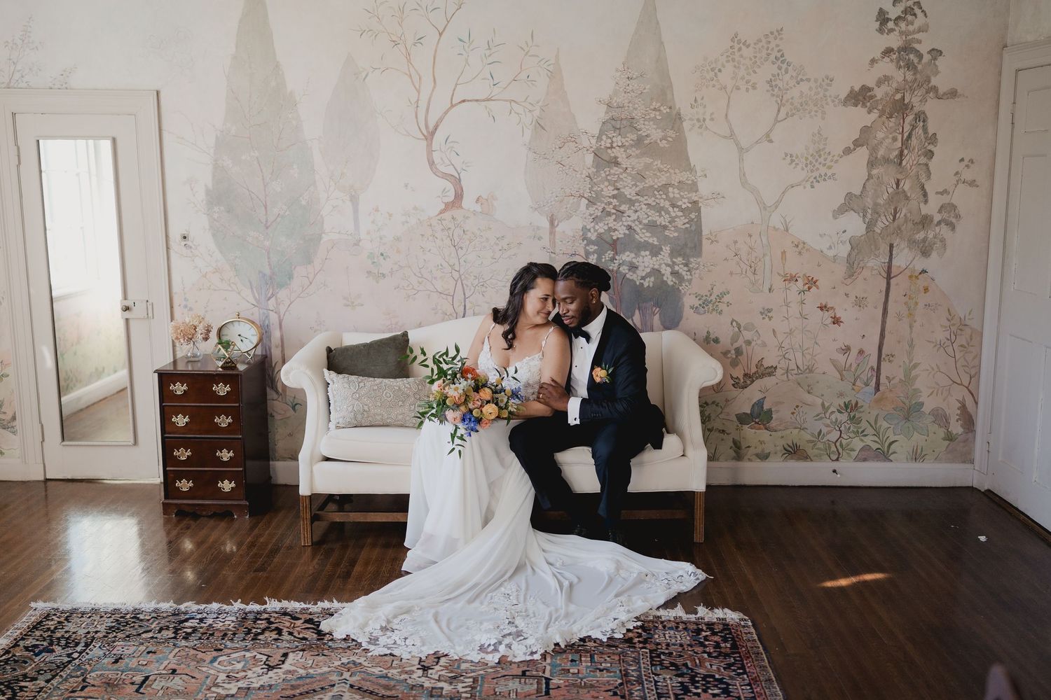 A wedding couple poses on an elegant stone balustrade balcony with architectural columns and greenery in the background.