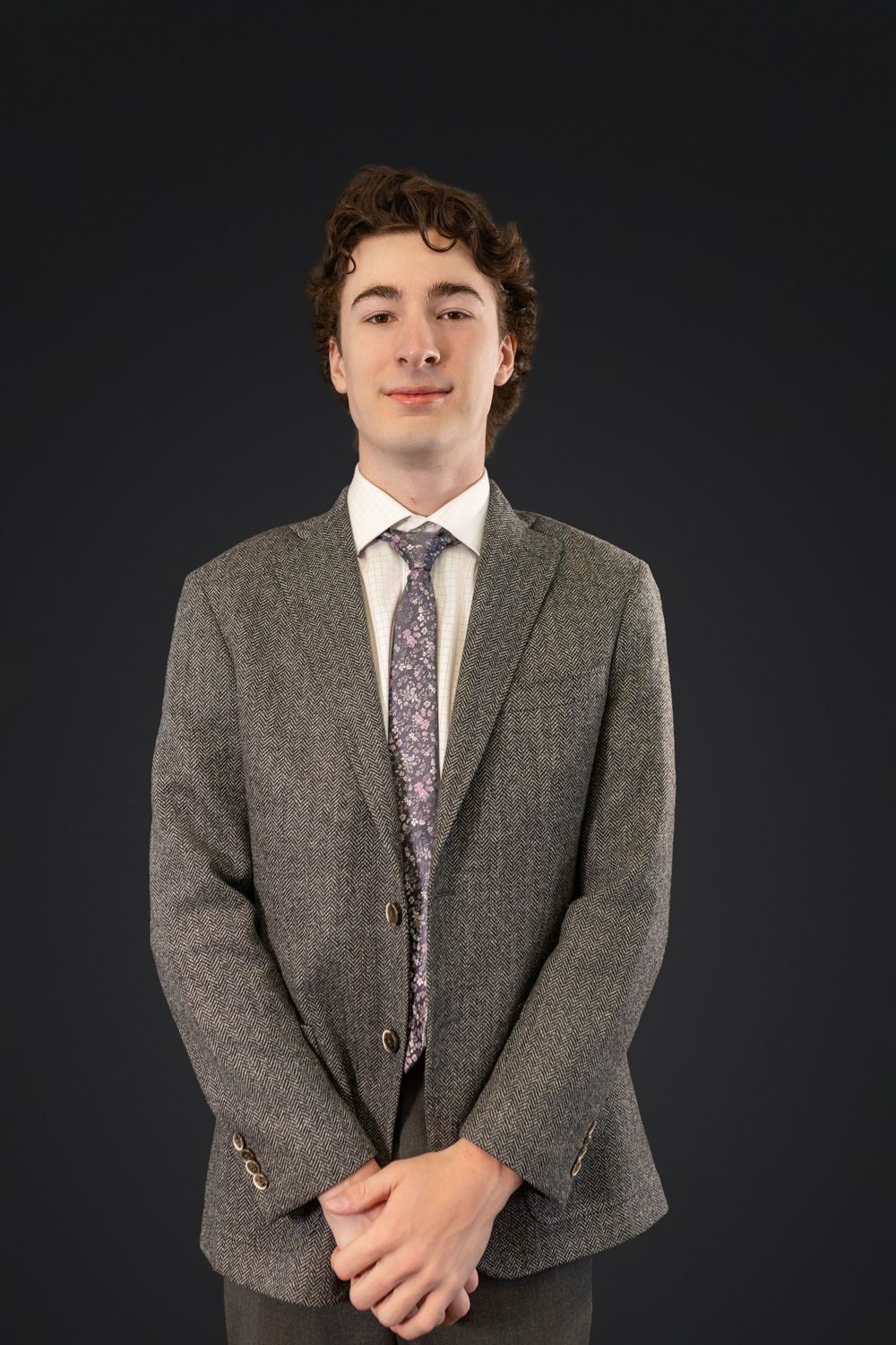Professional portrait of young person in grey suit jacket and floral tie against dark background.