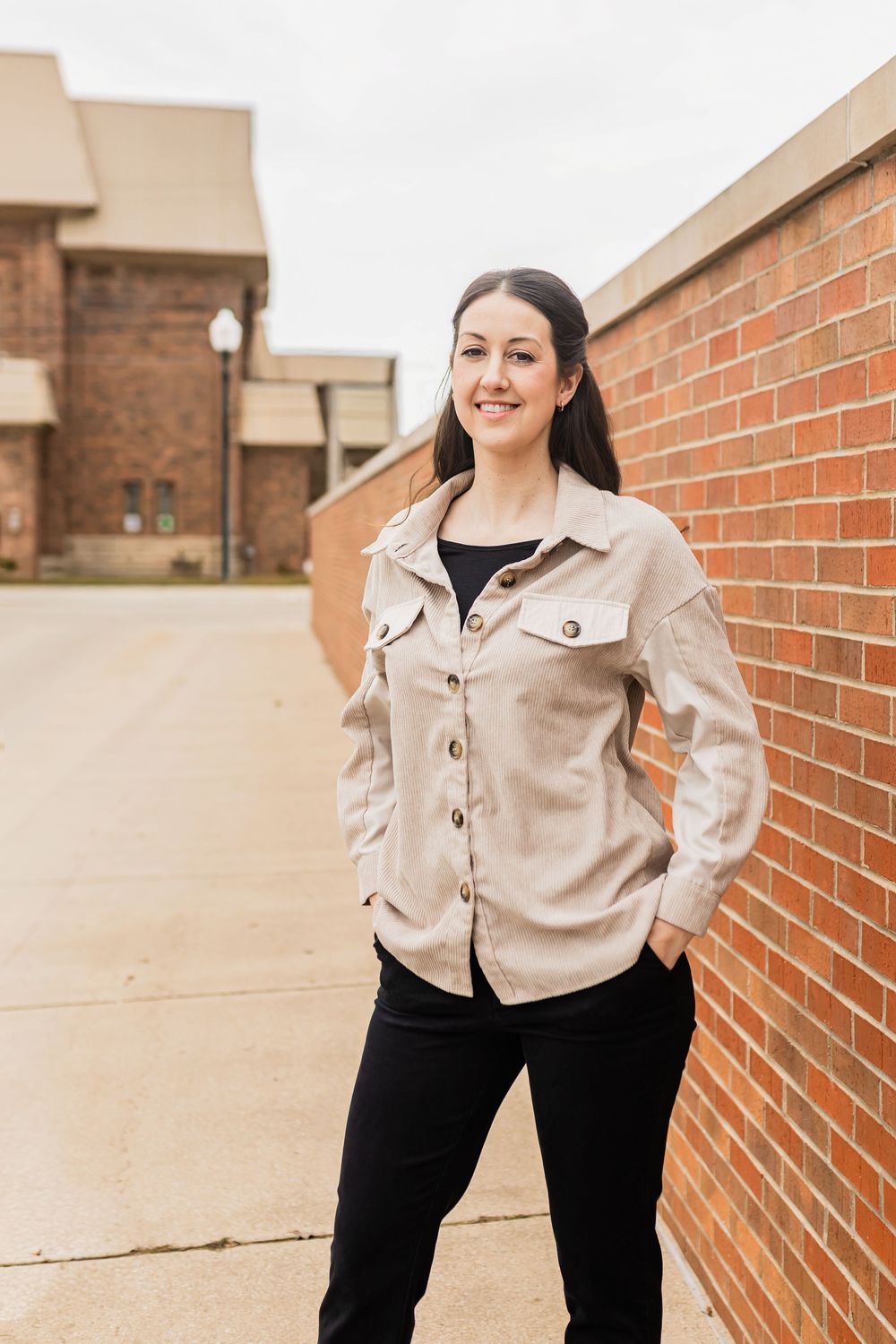 Young woman in beige jacket and black pants smiling outdoors near a brick wall.
