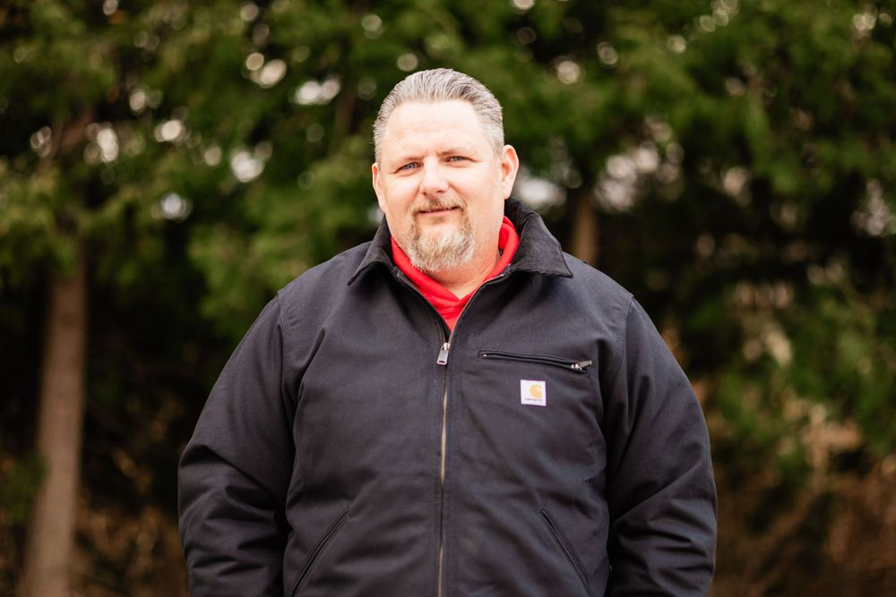 A person in a black Carhartt jacket and red shirt stands outdoors against a blurred natural background.