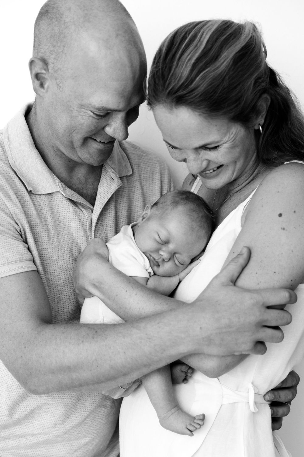 A tender black and white portrait of new parents embracing their sleeping newborn baby with loving smiles.