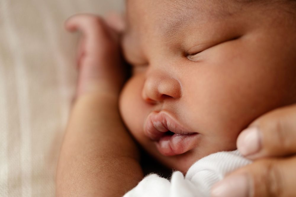 A sleeping newborn baby peacefully rests with parted lips against a soft cream-colored background.