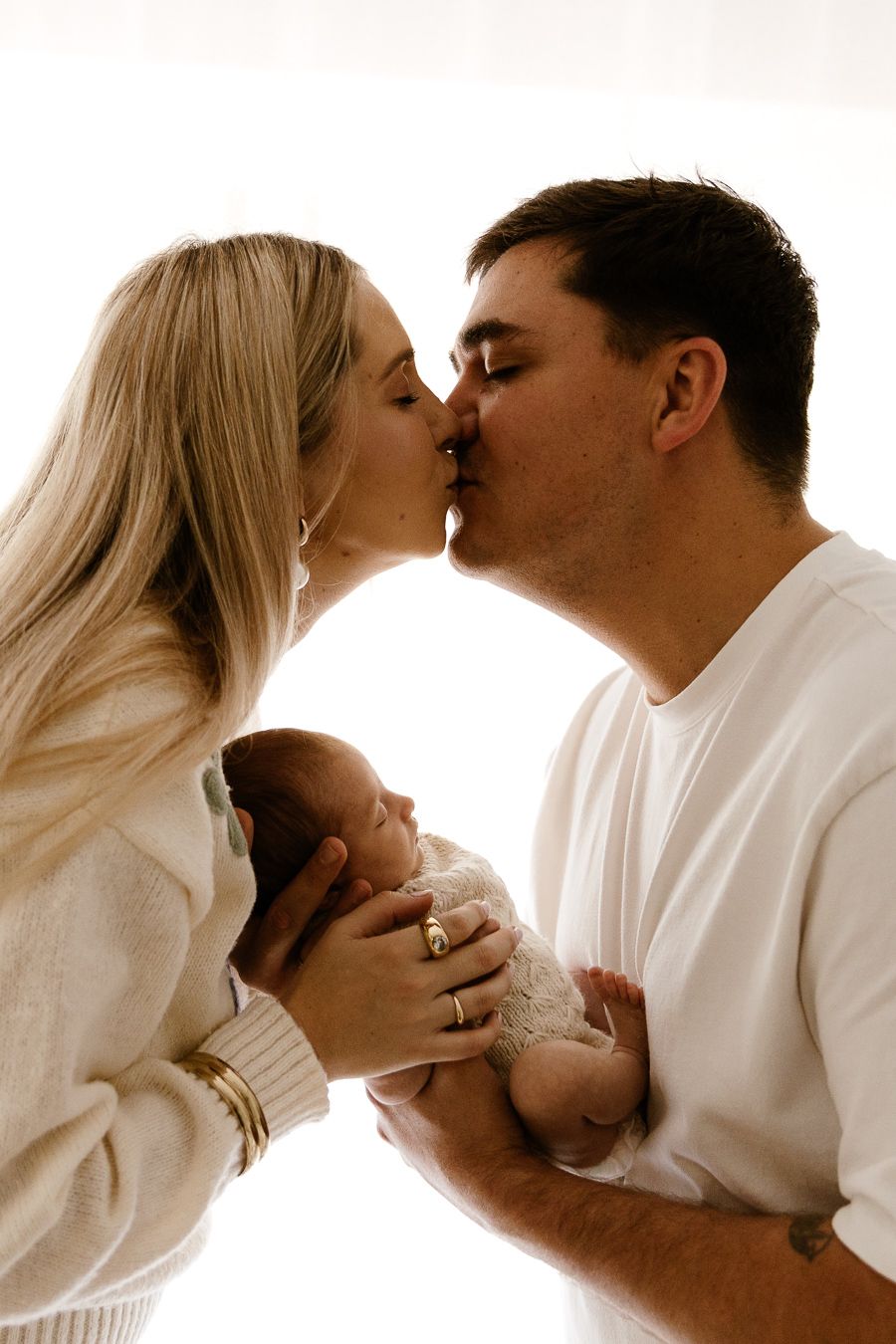 A couple shares a tender kiss while cradling their newborn baby in a bright, softly lit portrait.