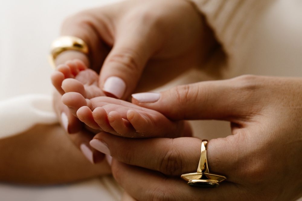 Adult hands gently holding a newborn baby's tiny feet, with gold rings visible on fingers.