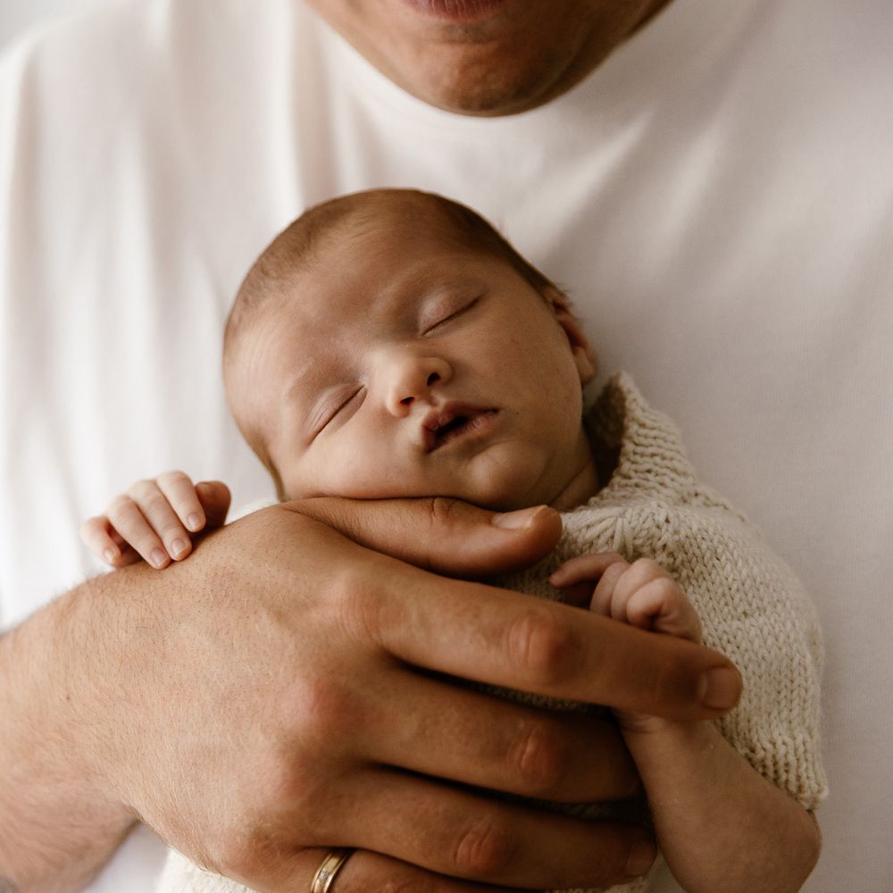 A peaceful newborn baby sleeping soundly in the secure arms of a parent wearing a white shirt and gold ring.