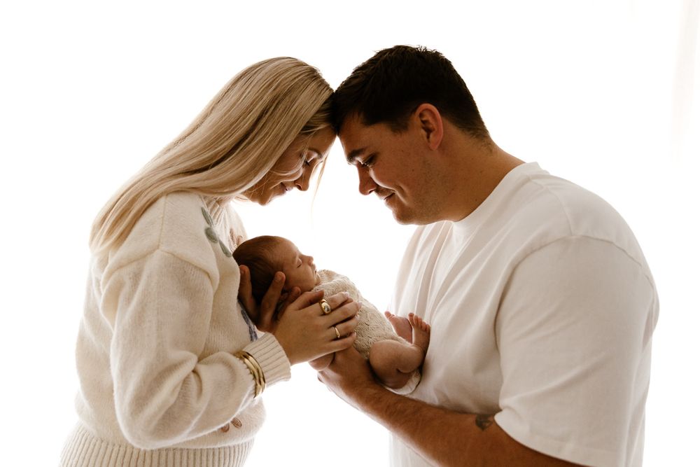New parents tenderly hold their newborn baby together, touching foreheads in an intimate moment against a white background.