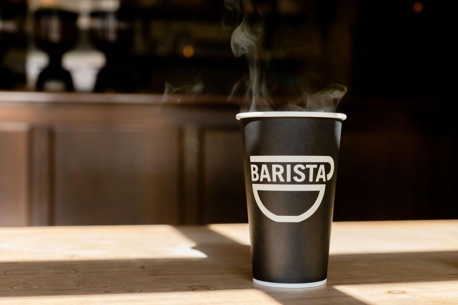 A steaming black disposable coffee cup with 'BARISTA' branding sits on a wooden counter in warm sunlight.