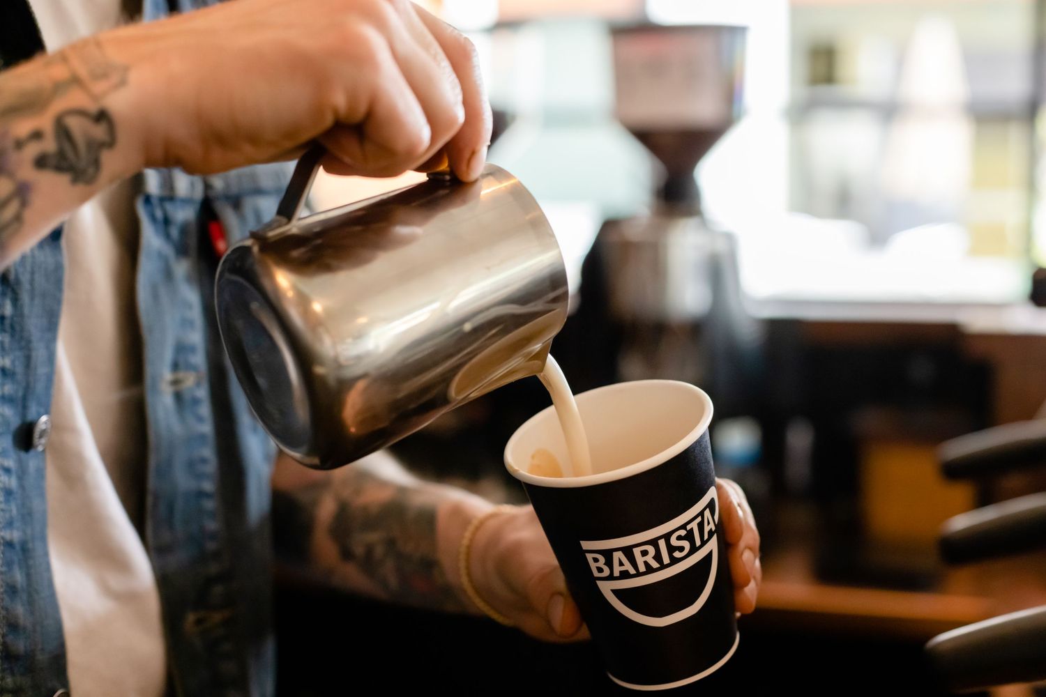 Steamed milk being poured from a metal pitcher into a black Bandit coffee cup on a cafe counter.