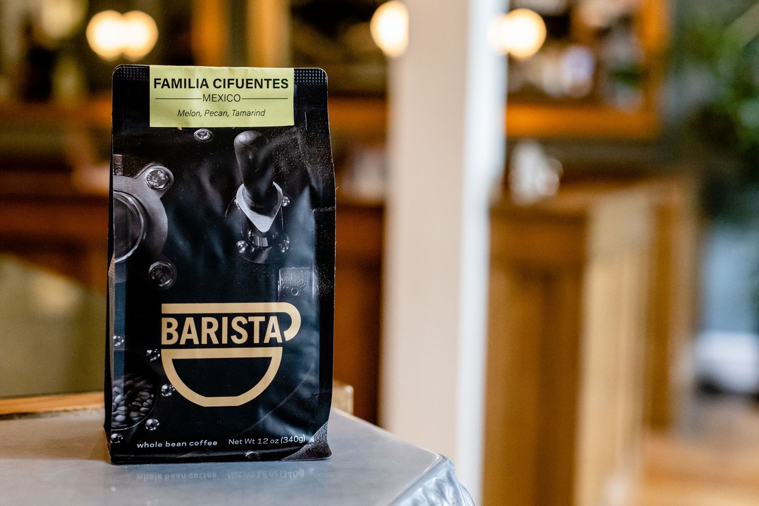 A black coffee bag labeled 'Barista' sits on a wooden surface with blurred warm lighting in the background.
