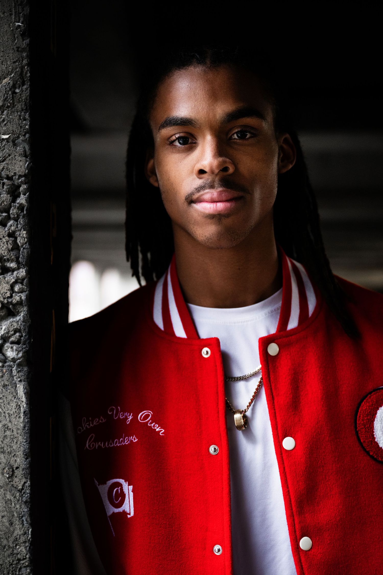 Urban portrait featuring person in red varsity jacket against dark textured wall.