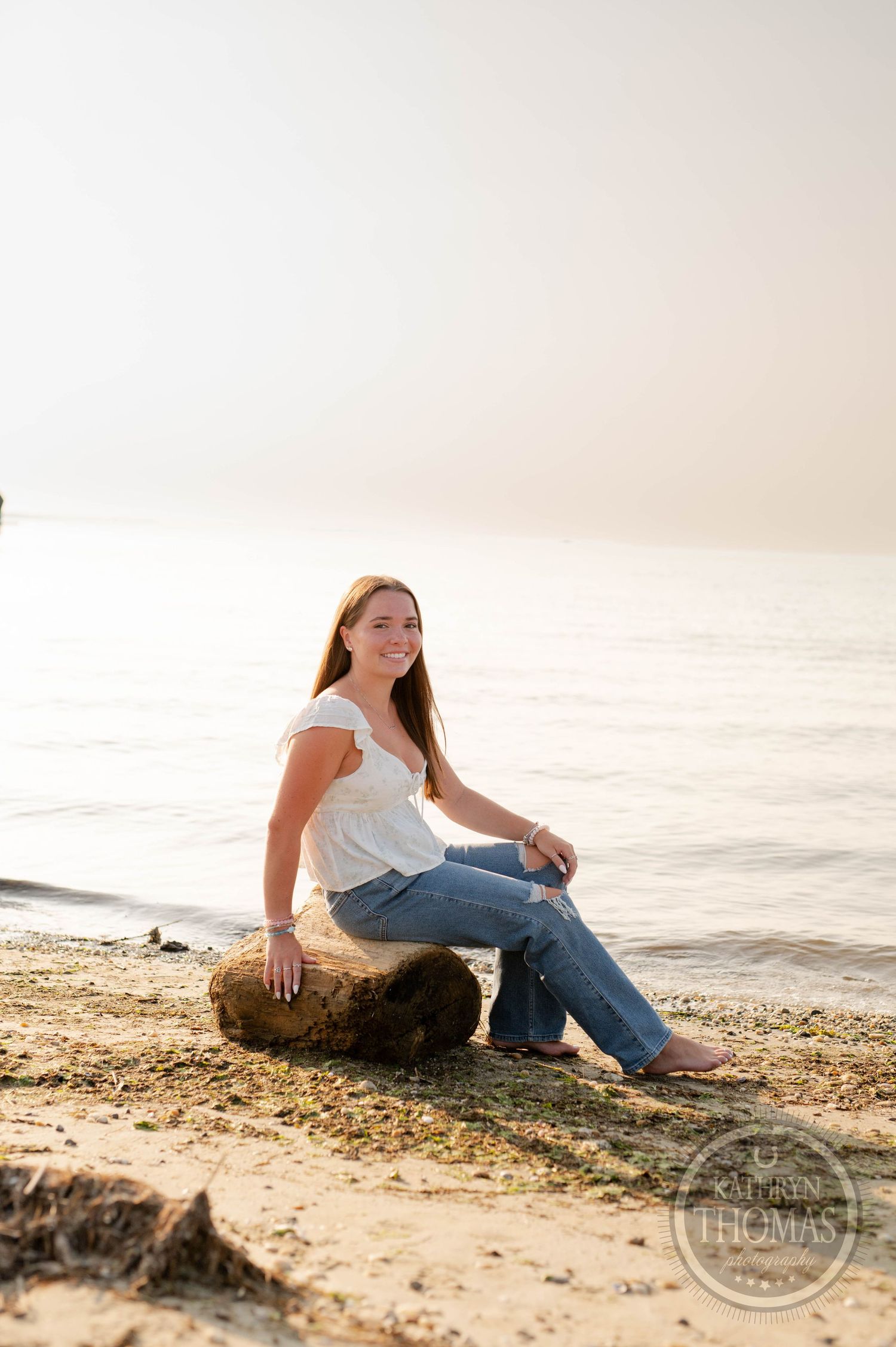 Sunset portrait of someone sitting on driftwood at the beach wearing jeans and a white top.