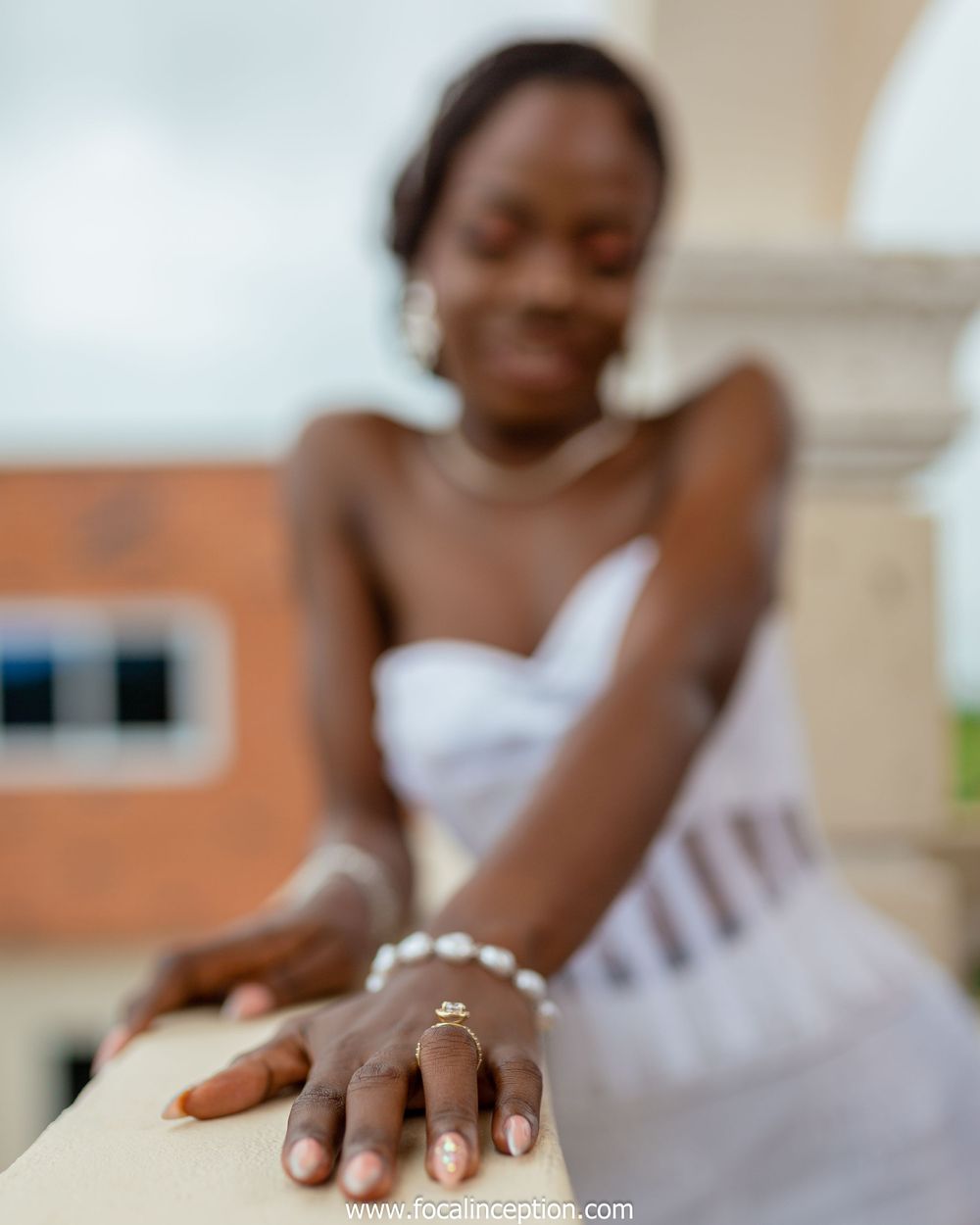 Close-up photo showing a pearl bracelet and engagement ring on a hand resting against white stone railing.