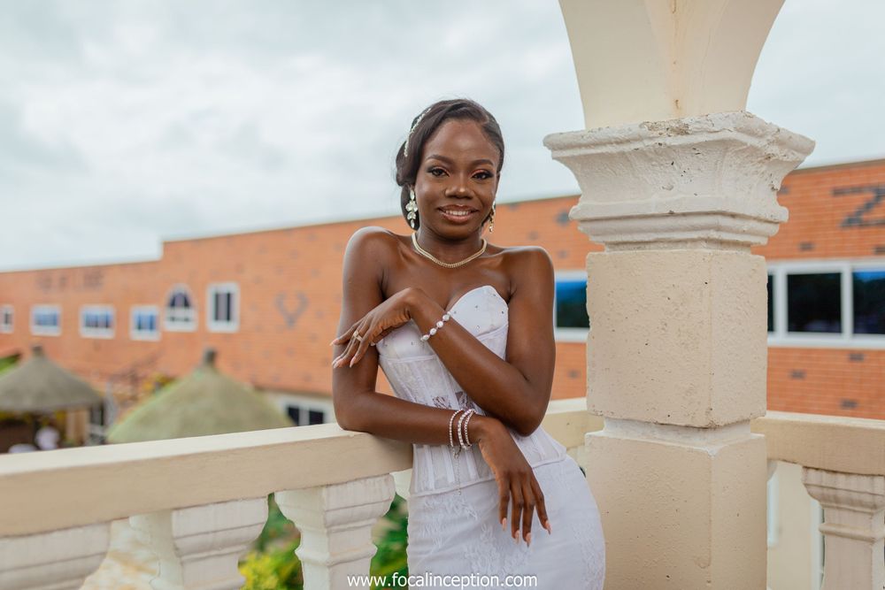 A person in an elegant white dress poses gracefully on a balcony with architectural columns and brick buildings in background.