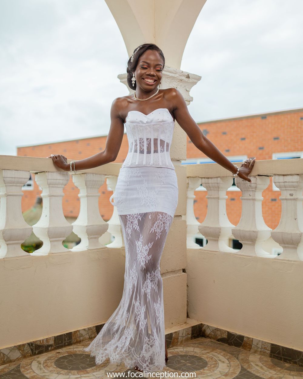 A woman wearing a stunning white lace wedding dress with corset detail poses elegantly on a balcony with ornate railings.