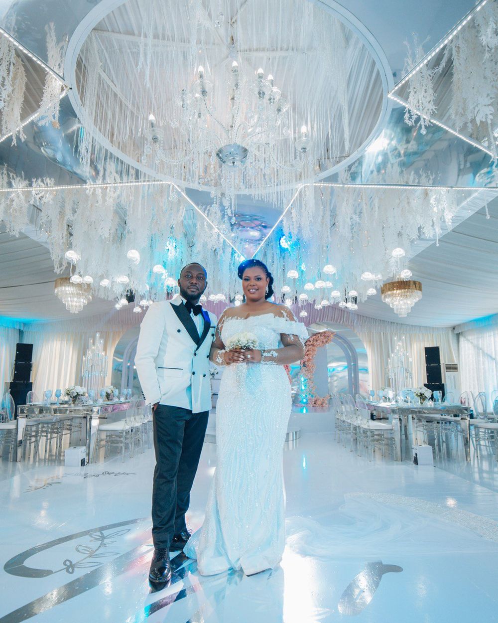 A wedding couple stands in an elegant white ballroom featuring crystal chandeliers and blue accent lighting.